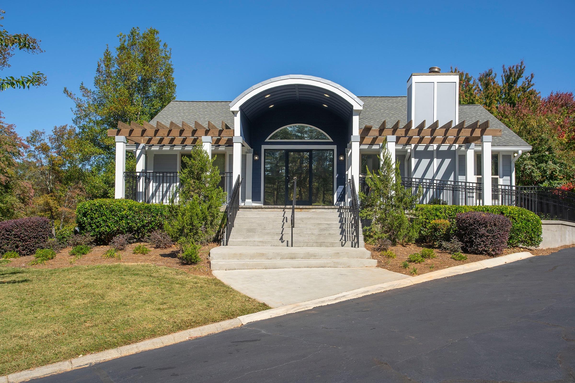 A modern building with a curved entrance, large front windows, and a flat roof. The exterior features white pillars and a wooden pergola. The surrounding landscape includes neatly trimmed bushes and grass, with a clear blue sky above. A paved pathway leads up to the entrance, set against a backdrop of trees.