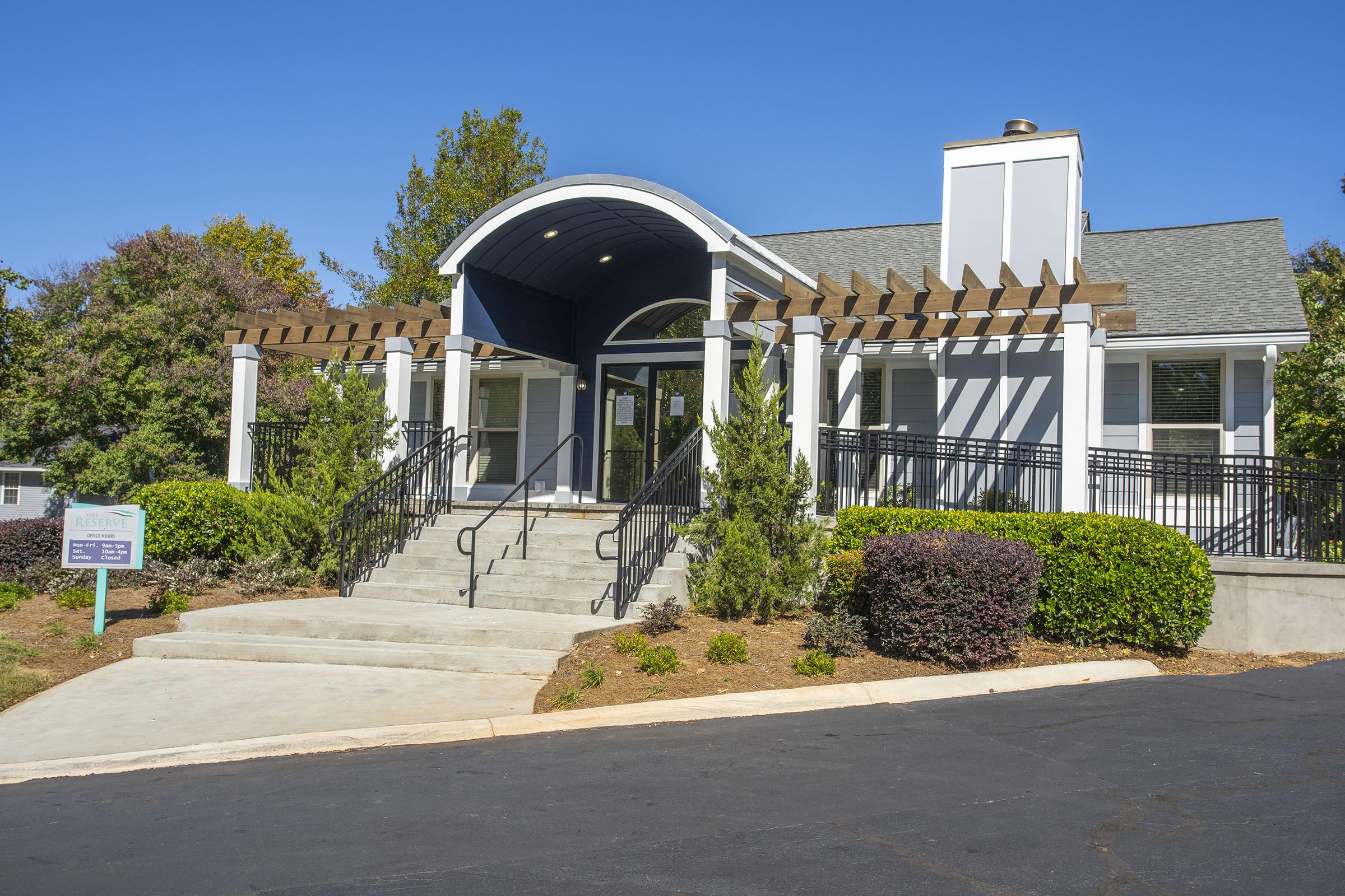 A modern building with a sloped roof and large entrance stairs. It features a white and blue exterior, surrounded by greenery and well-maintained landscaping. A sign is visible in front of the building, indicating its purpose or name. The sky is clear and blue, suggesting a sunny day.
