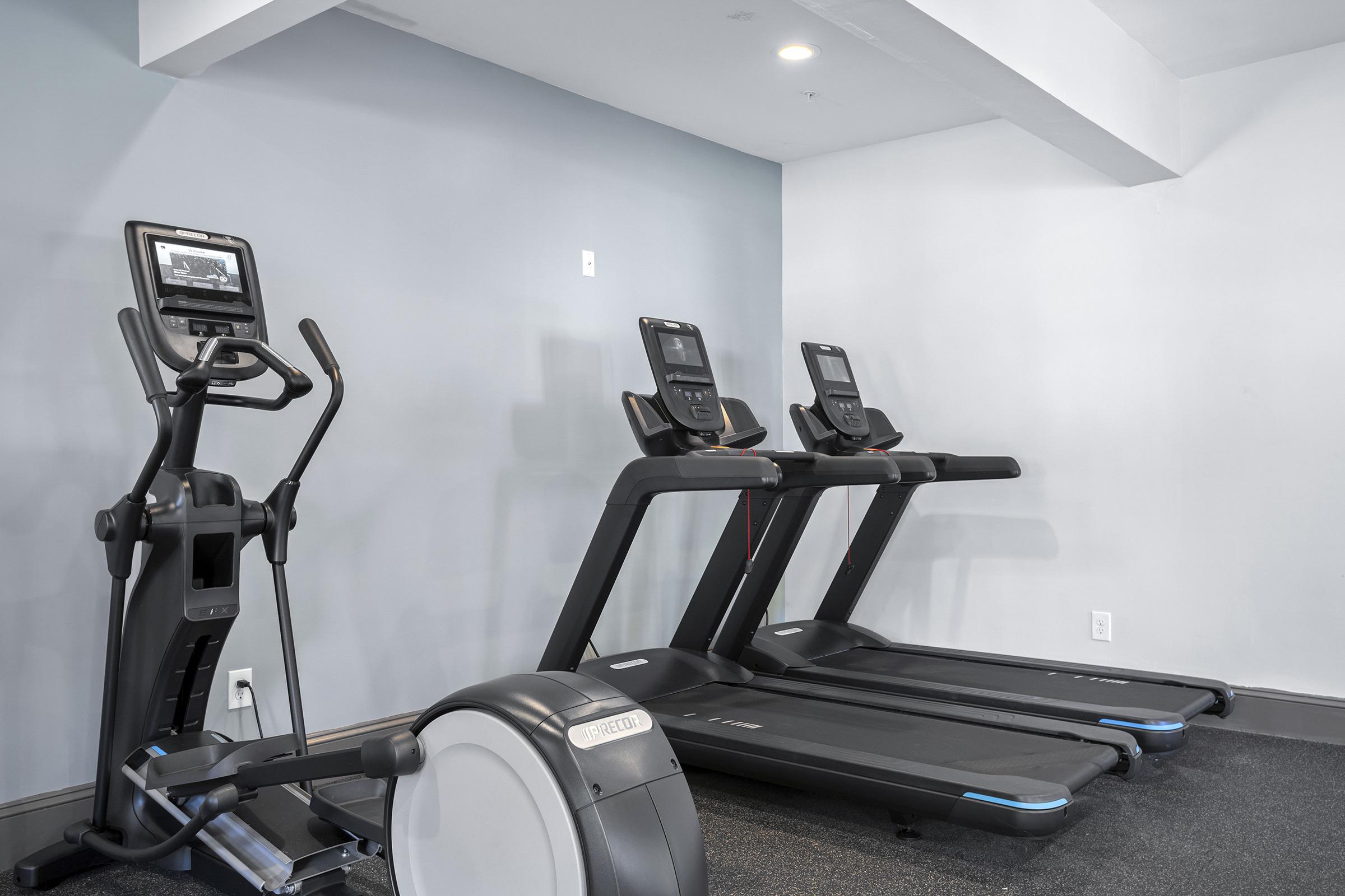 A fitness room featuring three treadmills aligned against a wall, alongside an elliptical machine. The walls are painted light grey, and the flooring is dark carpet, creating a modern and inviting workout space.