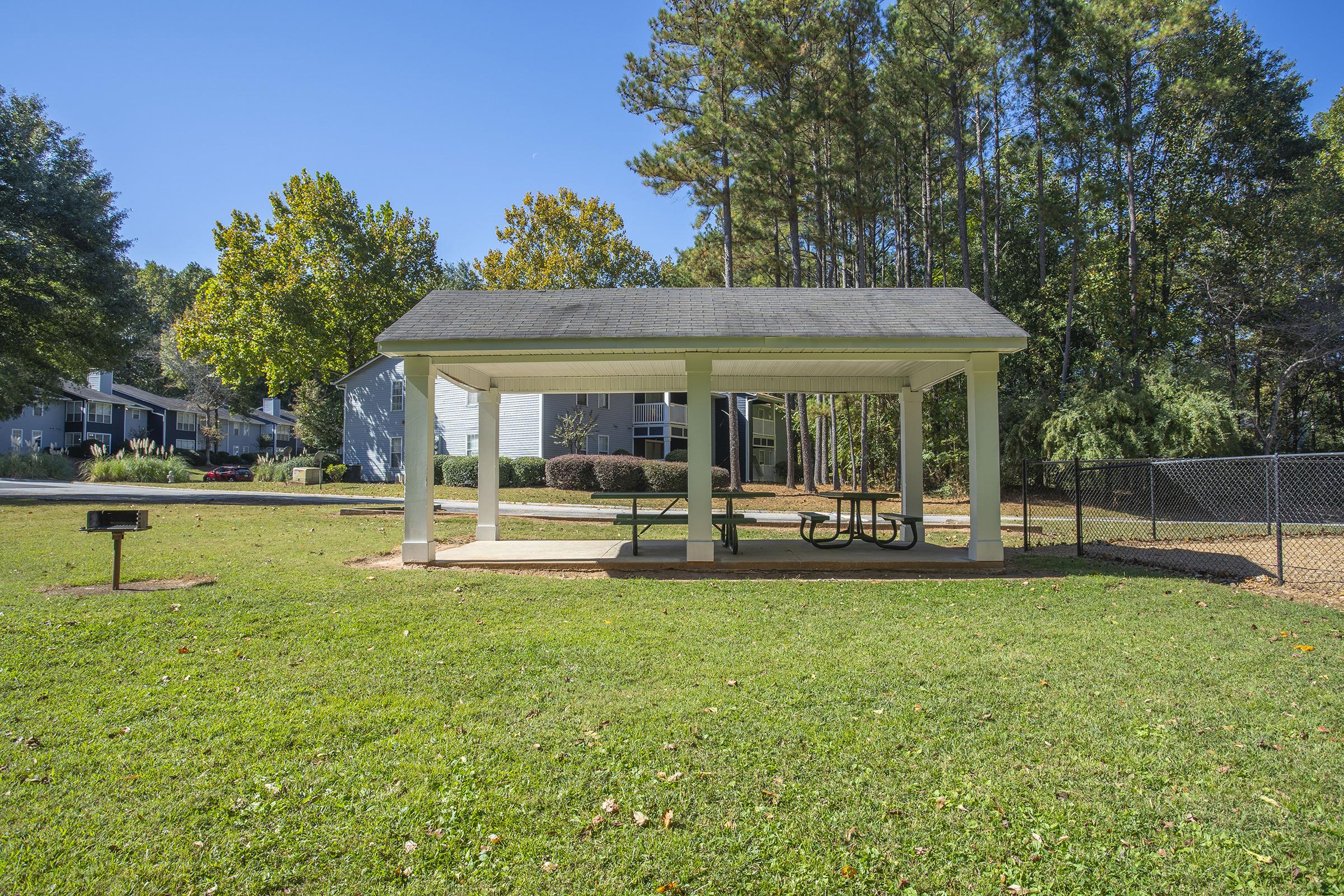 A covered picnic area with a picnic table and benches on a grassy patch, surrounded by trees. In the background, there are residential buildings and a chain-link fence. A charcoal grill is positioned nearby, and the sky is clear blue.