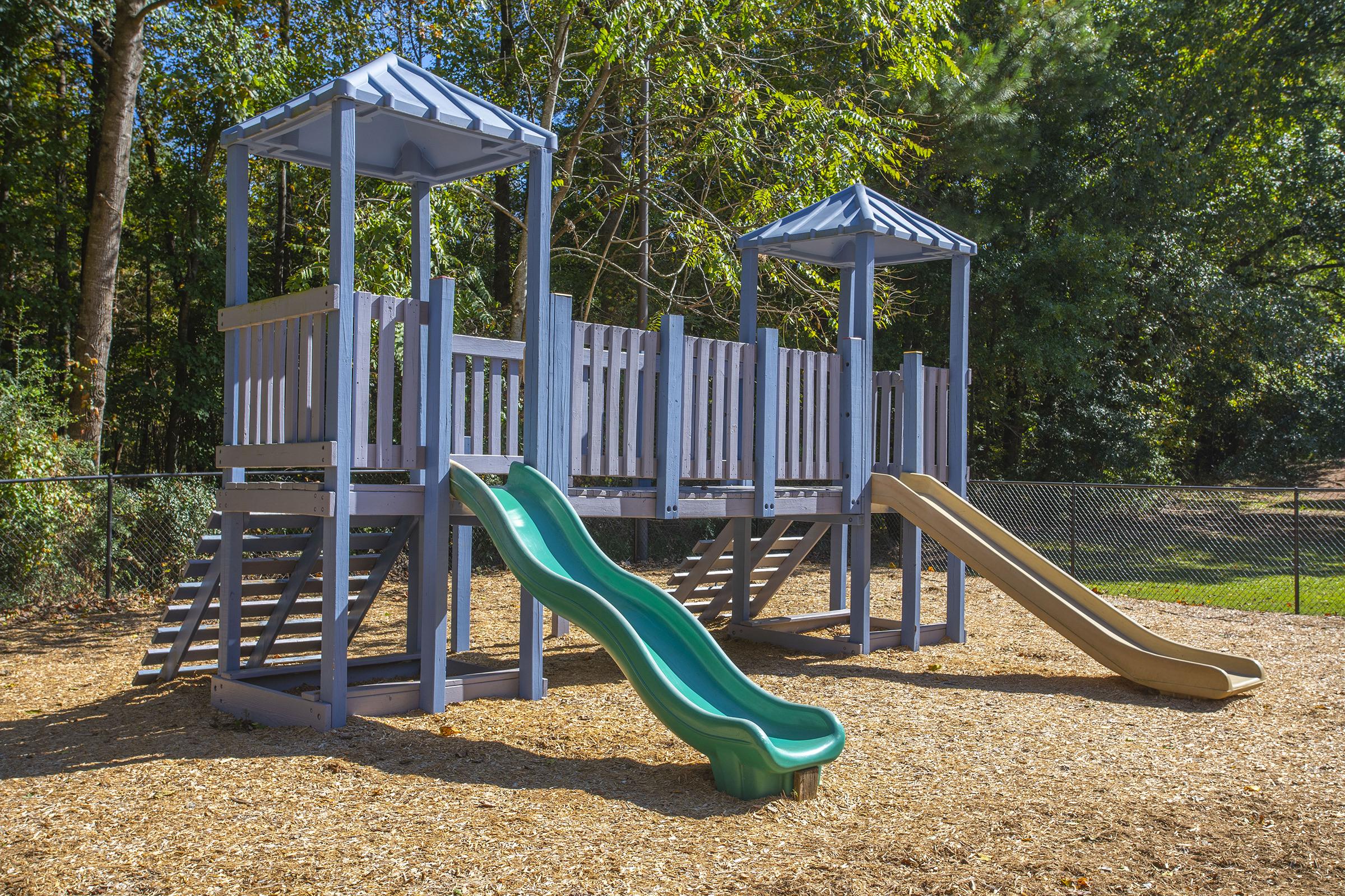A colorful playground structure featuring two slides—one green and one tan—set on a bed of wood chips. The structure has two raised play areas with roofs and ladders, surrounded by trees and a fenced area, offering a fun outdoor play environment for children.
