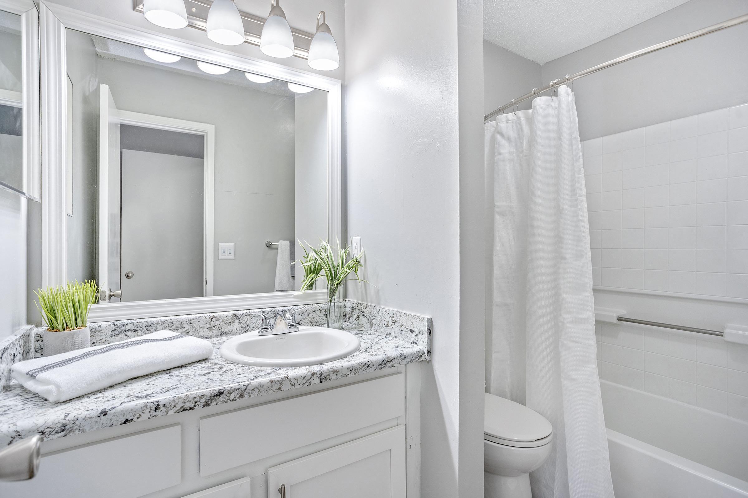 A bright, modern bathroom featuring a white vanity with a marble countertop, a round sink, and a neatly arranged towel. Above the sink, there are light fixtures. A shower with a white curtain is visible on the right, and the walls are painted in a soft gray color.