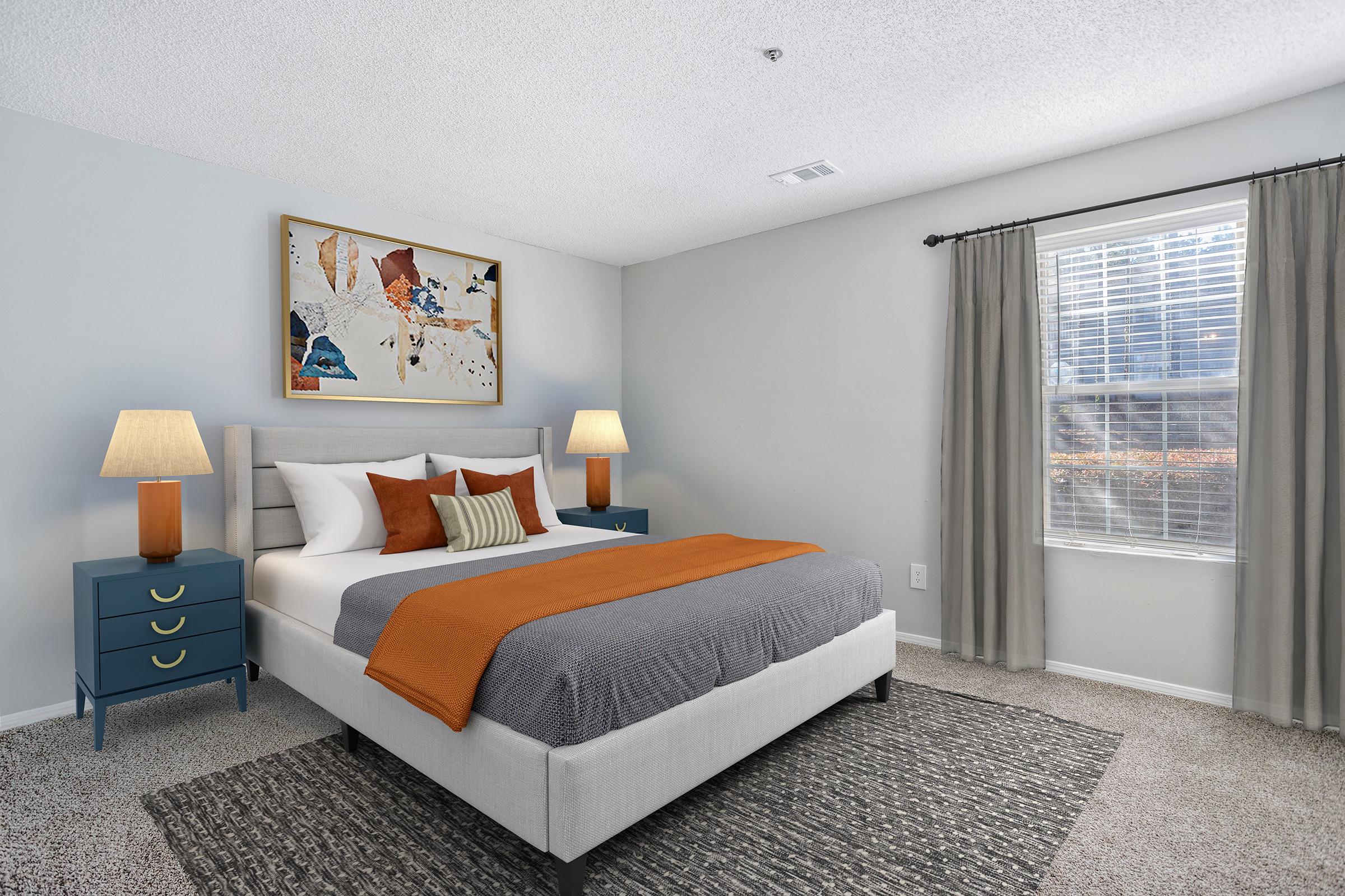 A well-decorated bedroom featuring a large bed with white bedding and an orange throw blanket. There are two bedside tables with lamps, a colorful abstract artwork on the wall, and a window with sheer curtains. The room has light gray walls and a soft carpet, creating a cozy ambiance.