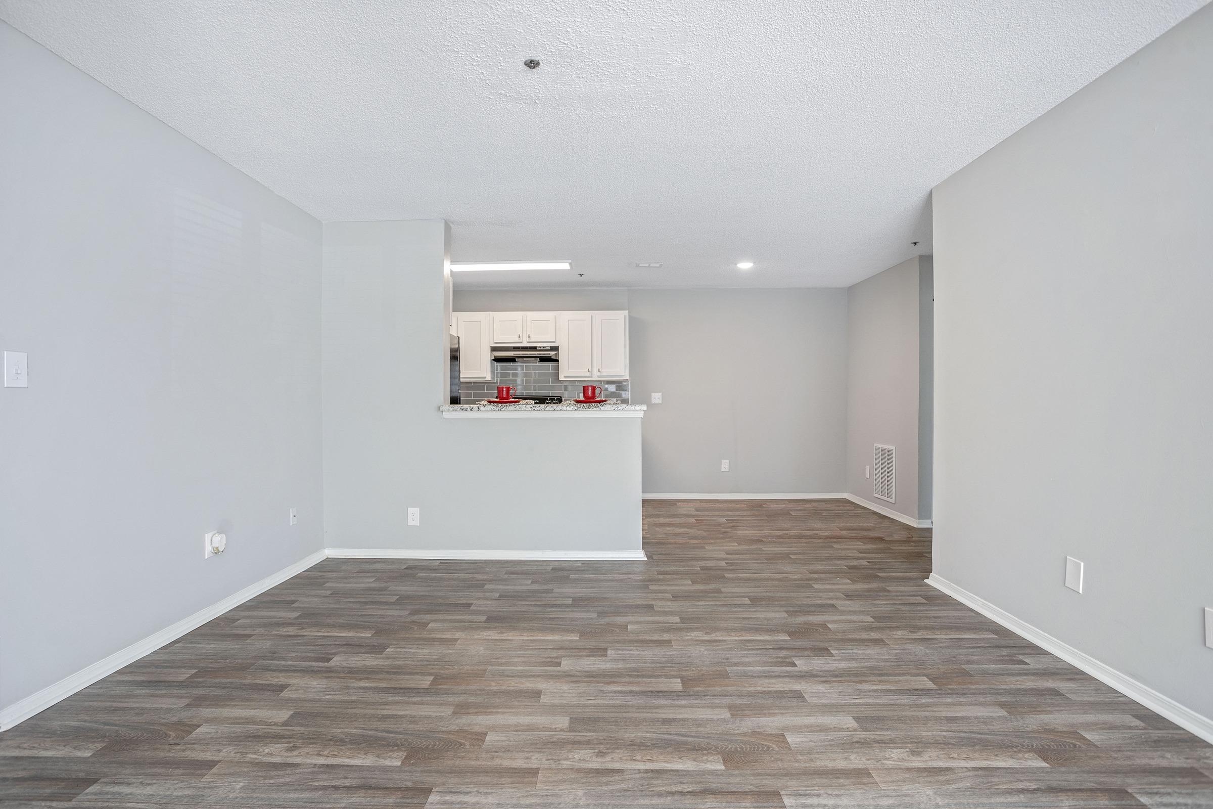 A spacious, empty living area with light gray walls and wood-style flooring. A kitchen is visible in the background featuring white cabinetry and a countertop. There are no furnishings, creating a clean and open atmosphere. Natural light illuminates the room through unseen windows.