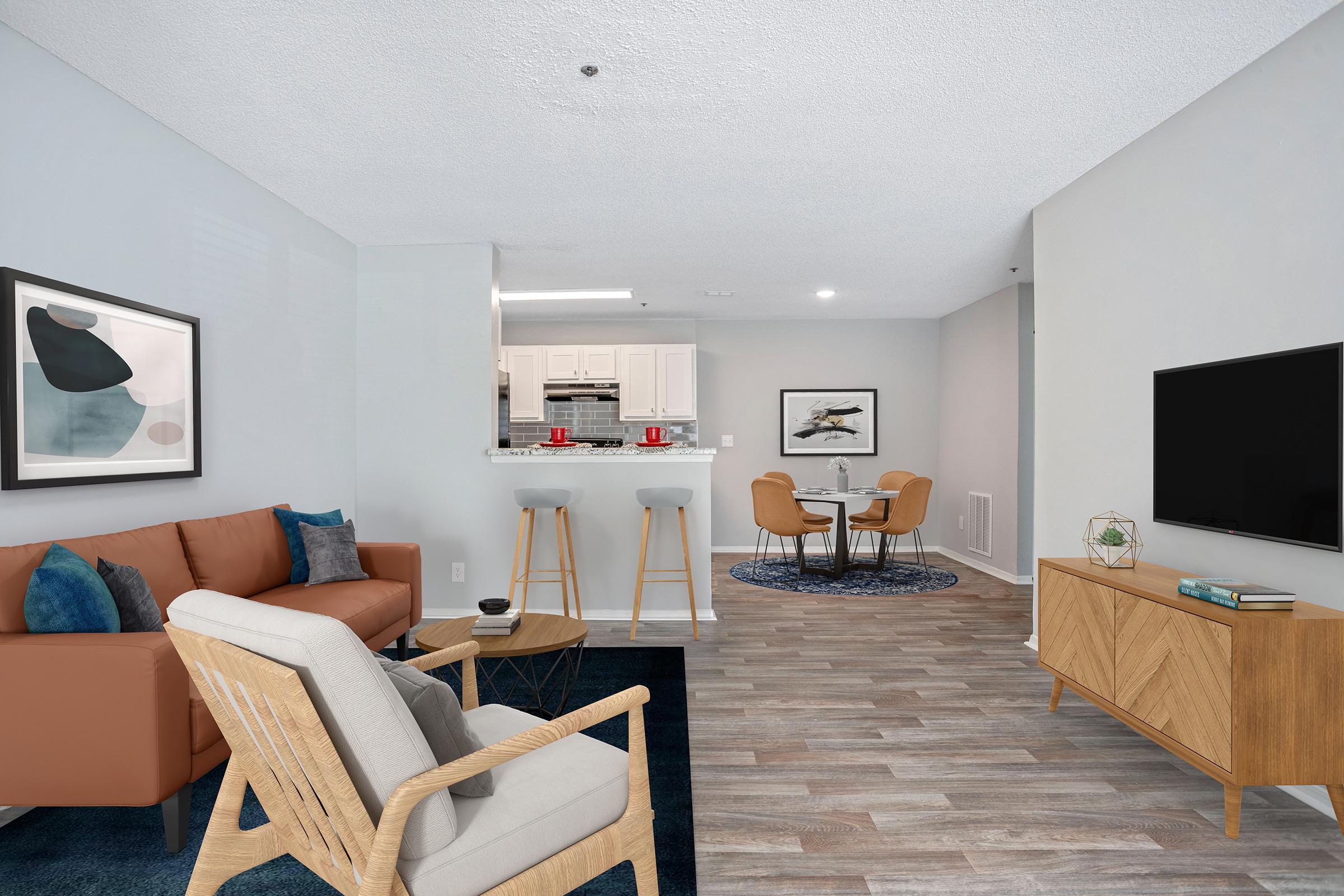Modern living room featuring a brown sofa, beige armchair, and wood side table. In the background, there's a dining area with a round table and two chairs. A sleek TV stand displays a TV, and a kitchen with white cabinets is visible. The floor has wooden tiles, and a decorative rug adds color.