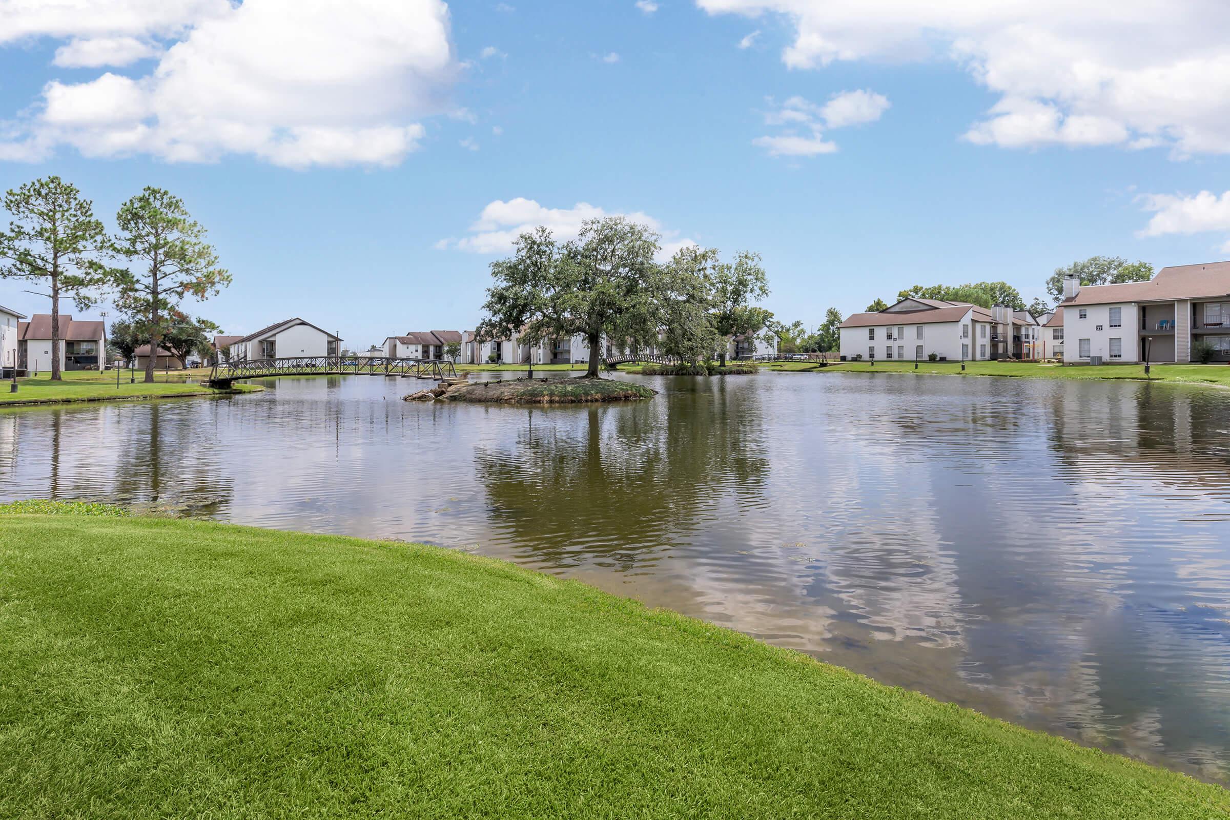 A peaceful lakeside scene featuring a tranquil body of water surrounded by lush green grass and trees. In the background, residential buildings reflect on the water's surface under a bright blue sky with scattered clouds. The setting conveys a serene, natural atmosphere.