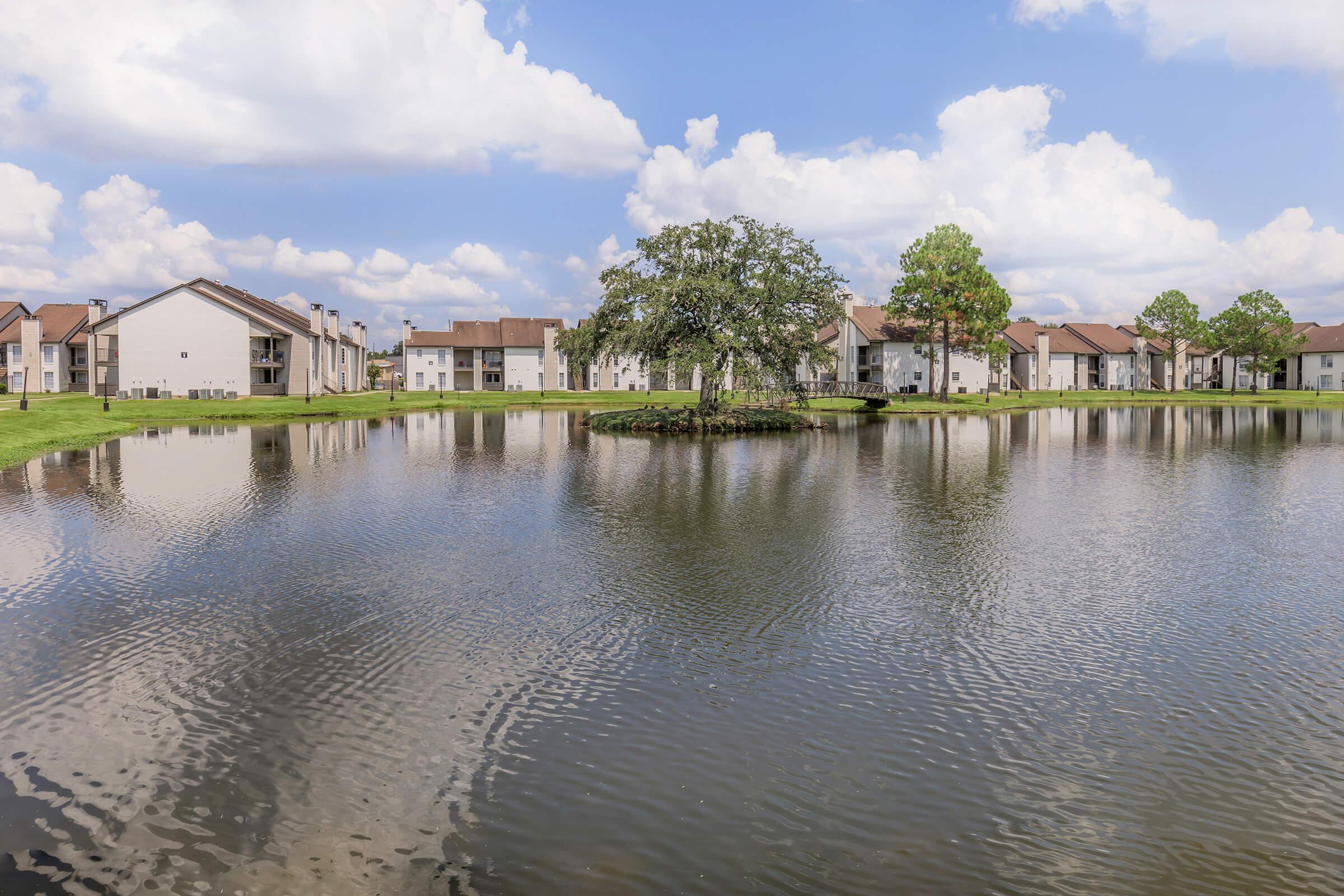 A serene view of a calm lake surrounded by residential buildings. In the center of the lake, there is a small island with a single tree. The sky is bright with fluffy white clouds, reflecting gently on the water's surface. Lush green grass borders the lake, creating a peaceful outdoor setting.