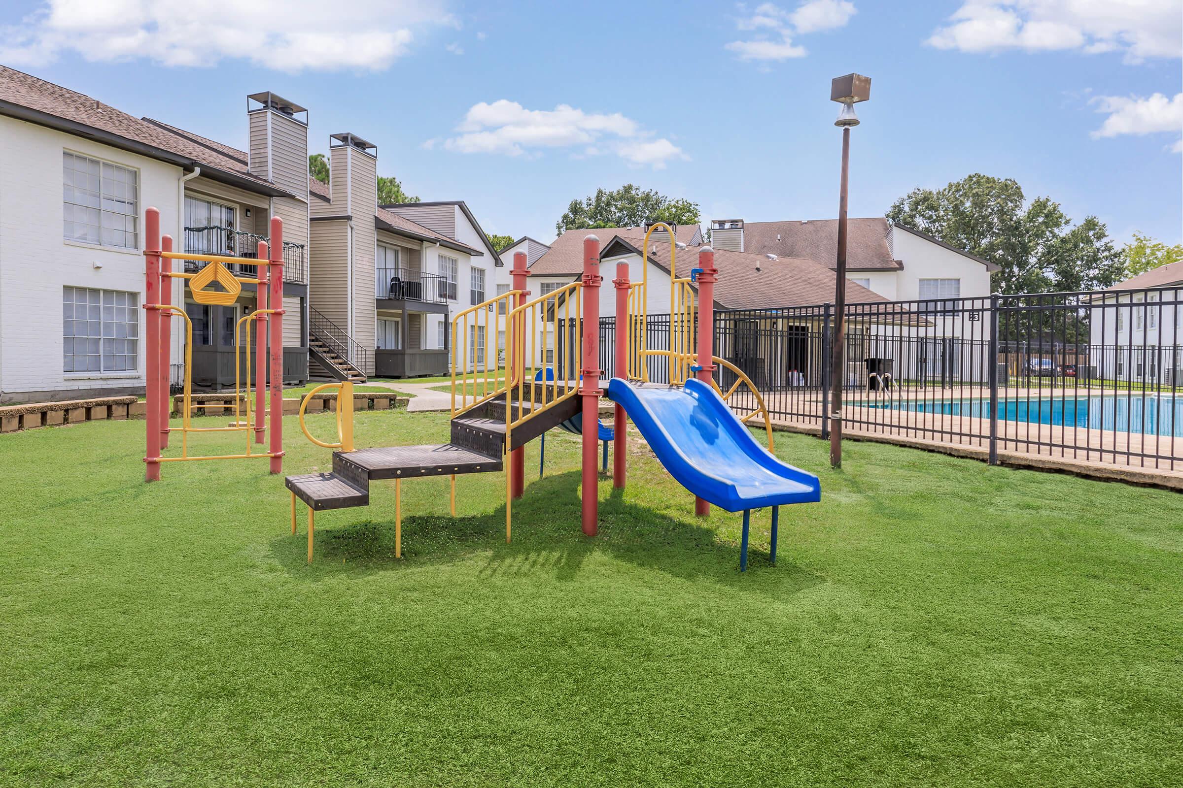 A brightly colored playground featuring a slide and climbing structures, situated on green artificial grass. In the background, there are apartment buildings and a fenced swimming pool. The sky is clear with a few clouds, creating a pleasant outdoor environment.