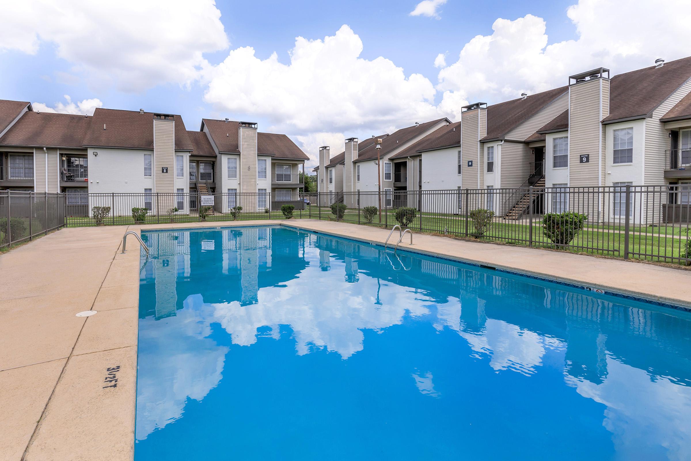 A serene swimming pool surrounded by an apartment complex, with clear blue water reflecting the sky and clouds. The pool area is fenced and features neatly trimmed bushes and lounging spaces. The buildings have a simple architectural design, creating a peaceful residential atmosphere.
