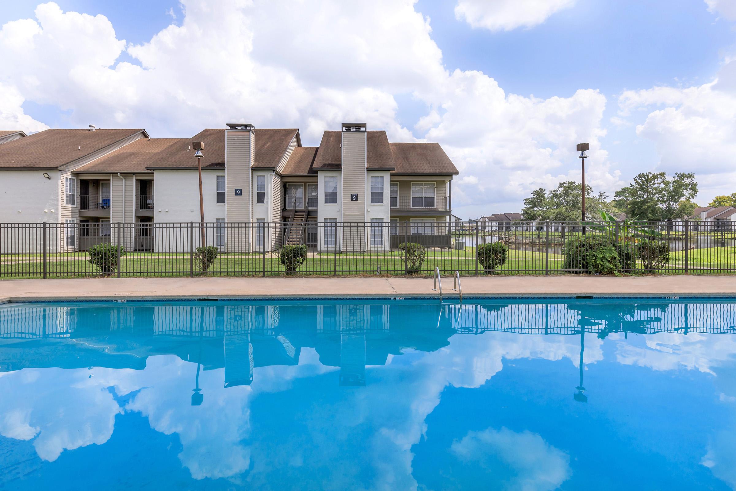 A serene view of a sparkling blue swimming pool reflecting clouds and nearby buildings. The pool area is surrounded by a black fence and lush green grass, with two-story apartment buildings in the background under a partly cloudy sky.