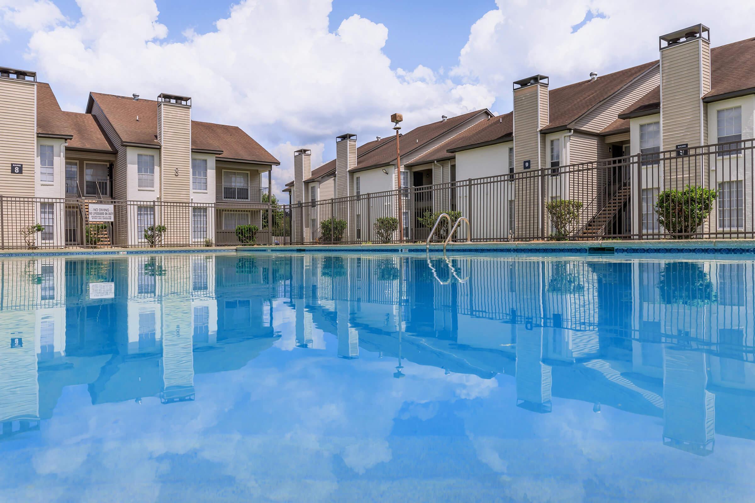 A clear swimming pool reflecting the sky and clouds, surrounded by a fenced area and several residential buildings with brown roofs. Lush greenery is visible along the edges of the pool area.