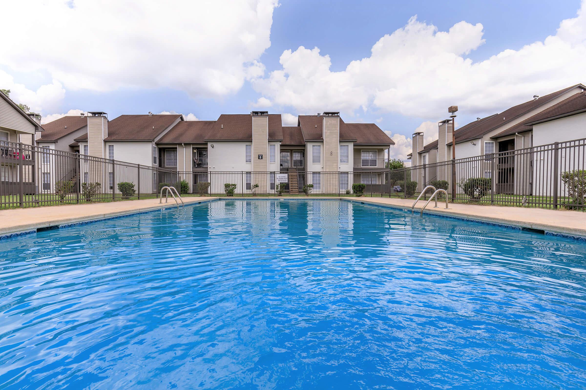 A view of a clean, blue swimming pool surrounded by well-maintained grass and shrubbery. In the background, there are residential buildings with balconies under a partly cloudy sky. The pool area is enclosed by a decorative fence, creating a relaxing outdoor space.