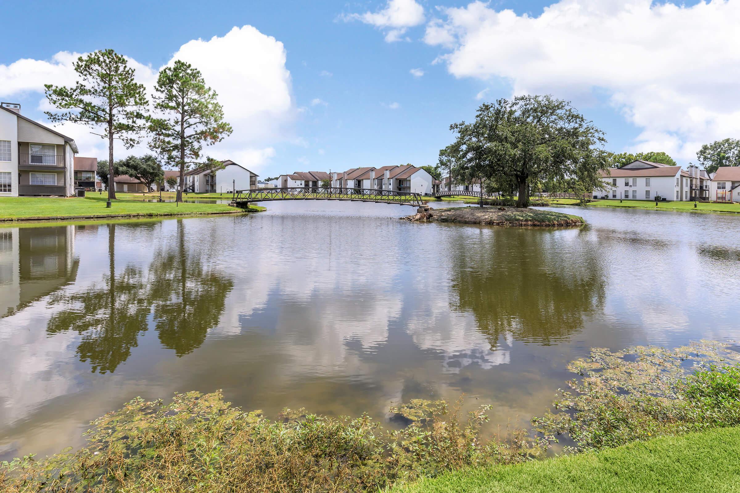 A serene pond surrounded by lush greenery and residential buildings. The calm water reflects the blue sky and white clouds, while a small island with a tree adds a natural focal point. A wooden bridge spans the pond, enhancing the picturesque landscape.