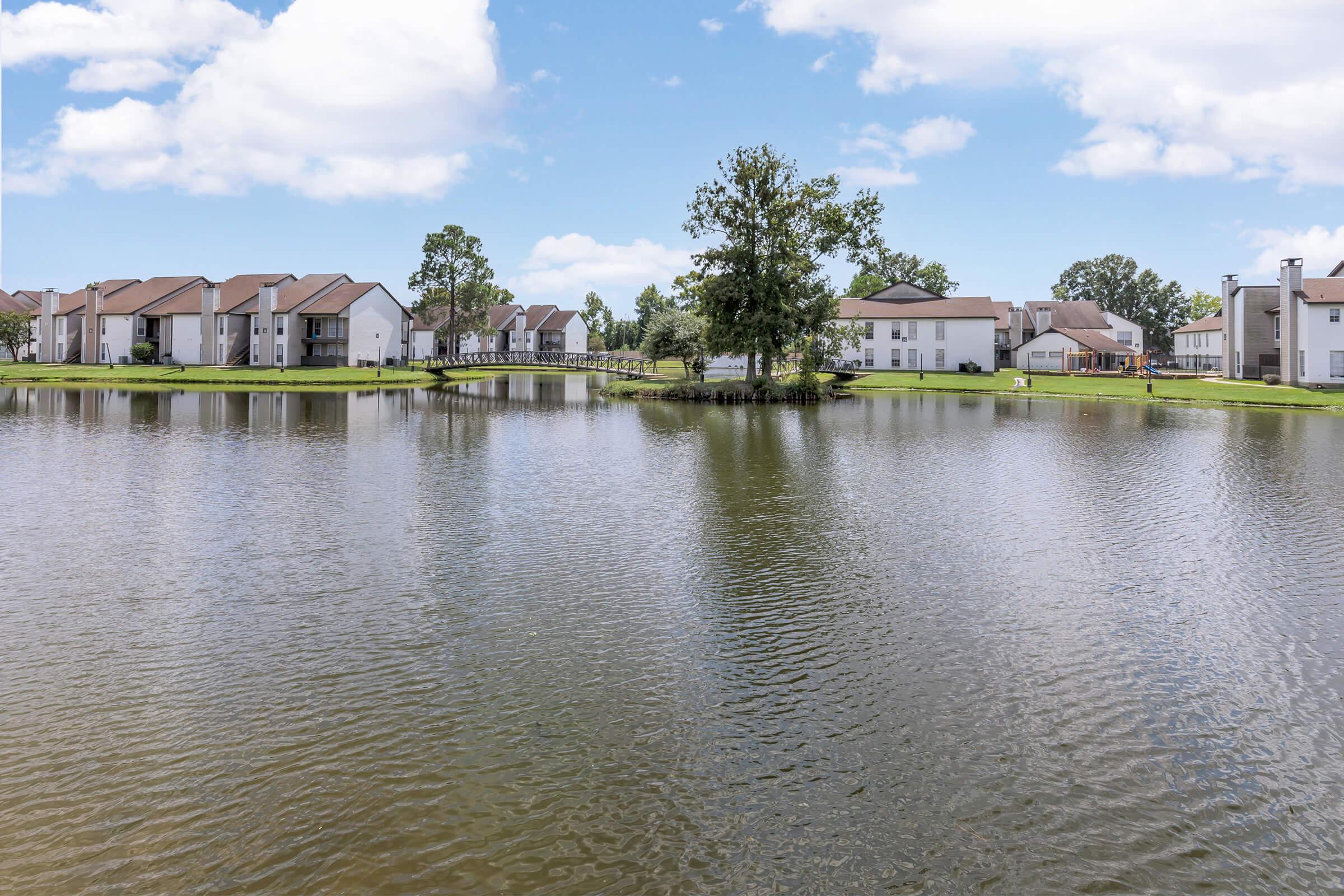 A tranquil pond surrounded by residential buildings, with lush green trees and a small bridge in the background. The calm water reflects the blue sky and fluffy clouds, creating a serene atmosphere in a suburban setting.