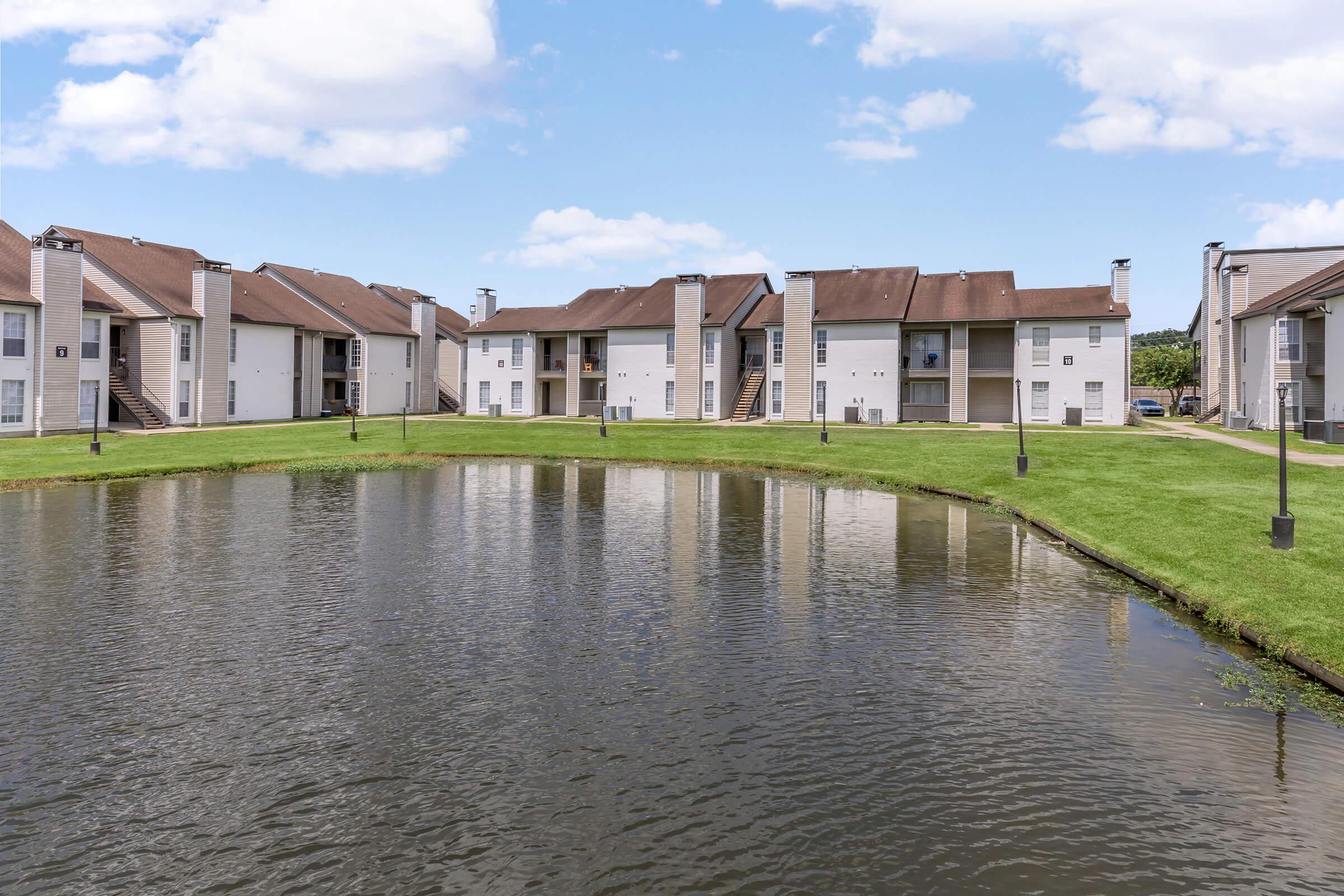 View of a multi-unit residential complex with two-story buildings, set beside a tranquil pond. The landscape features well-maintained grass and a clear sky with a few clouds, creating a peaceful suburban atmosphere.