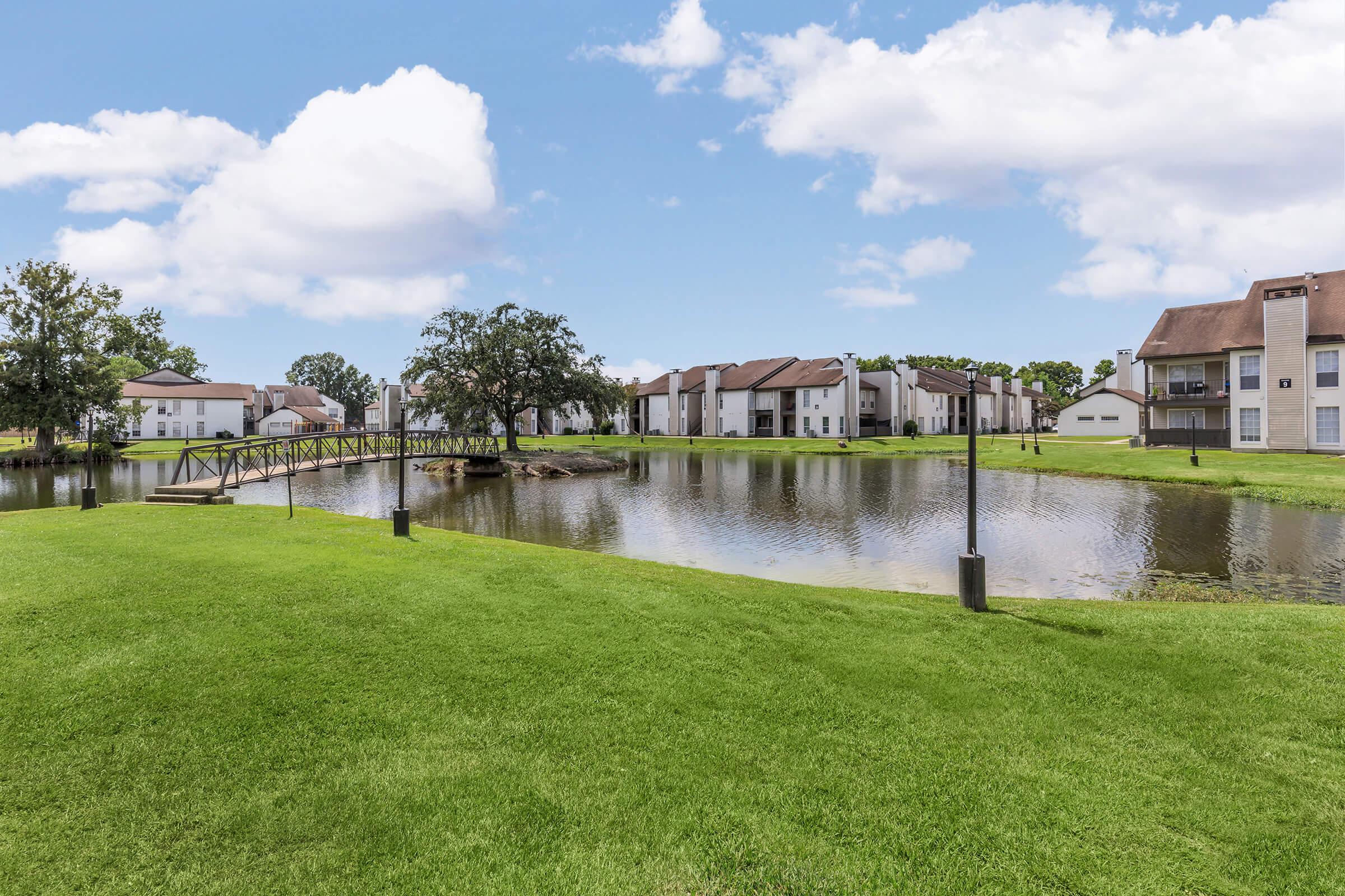 View of a well-maintained residential area featuring a pond, grassy landscape, and a wooden footbridge. The background includes several apartment buildings under a partly cloudy sky, creating a serene and inviting atmosphere.