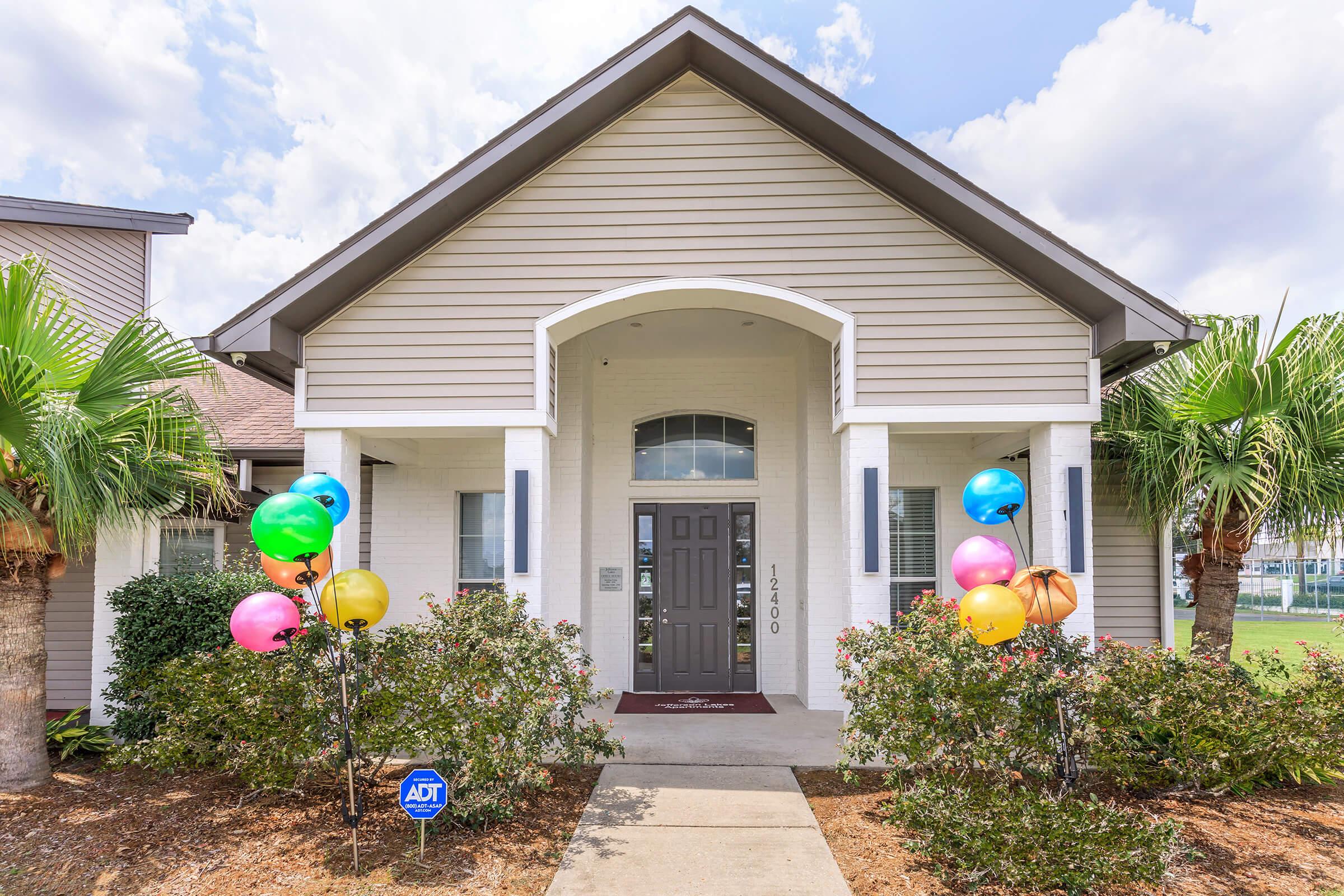 A welcoming entrance to a building featuring a gray front door and large windows. Colorful balloons in various shades adorn the porch area. Lush palm trees and shrubbery line the path leading to the entrance, set against a partly cloudy blue sky.