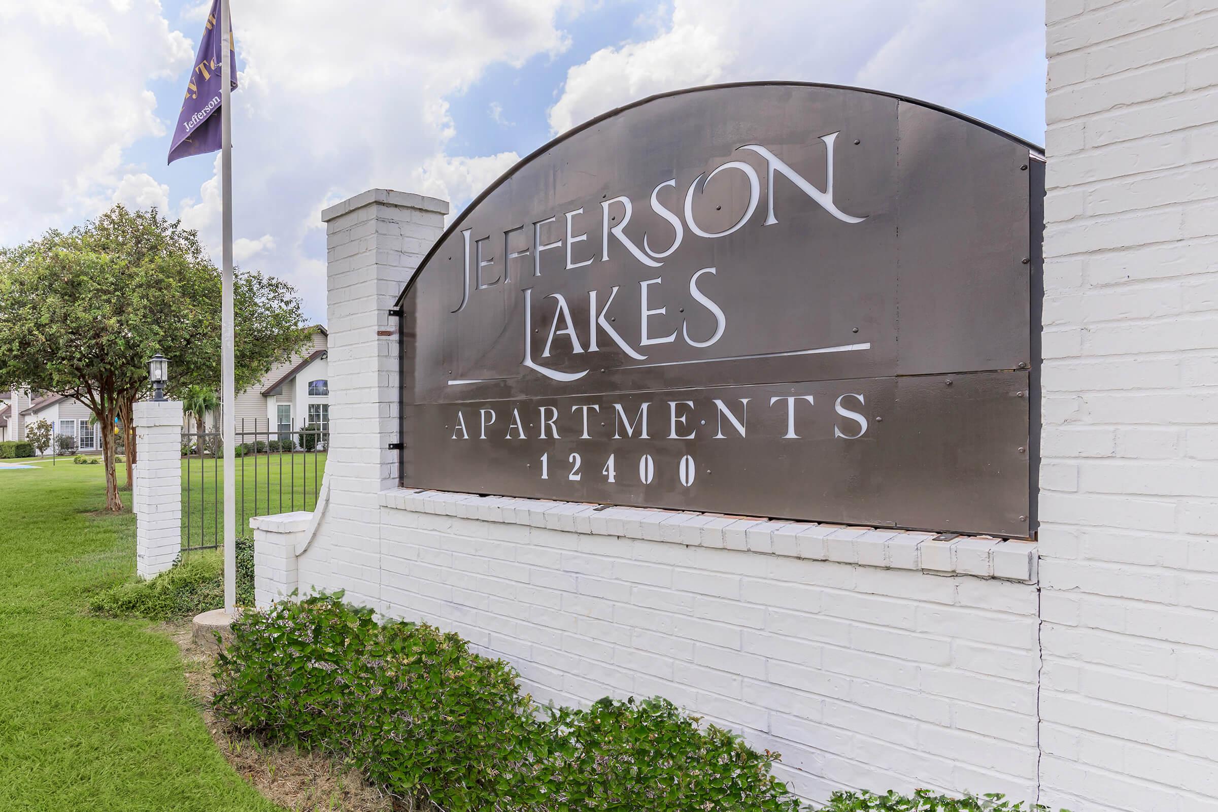 Sign for Jefferson Lakes Apartments, featuring the name prominently displayed on a black background with white lettering. The sign is situated near green grass and shrubs, with a flagpole in the background indicating a nearby flag. The structure has a white brick finish and is part of an apartment complex.