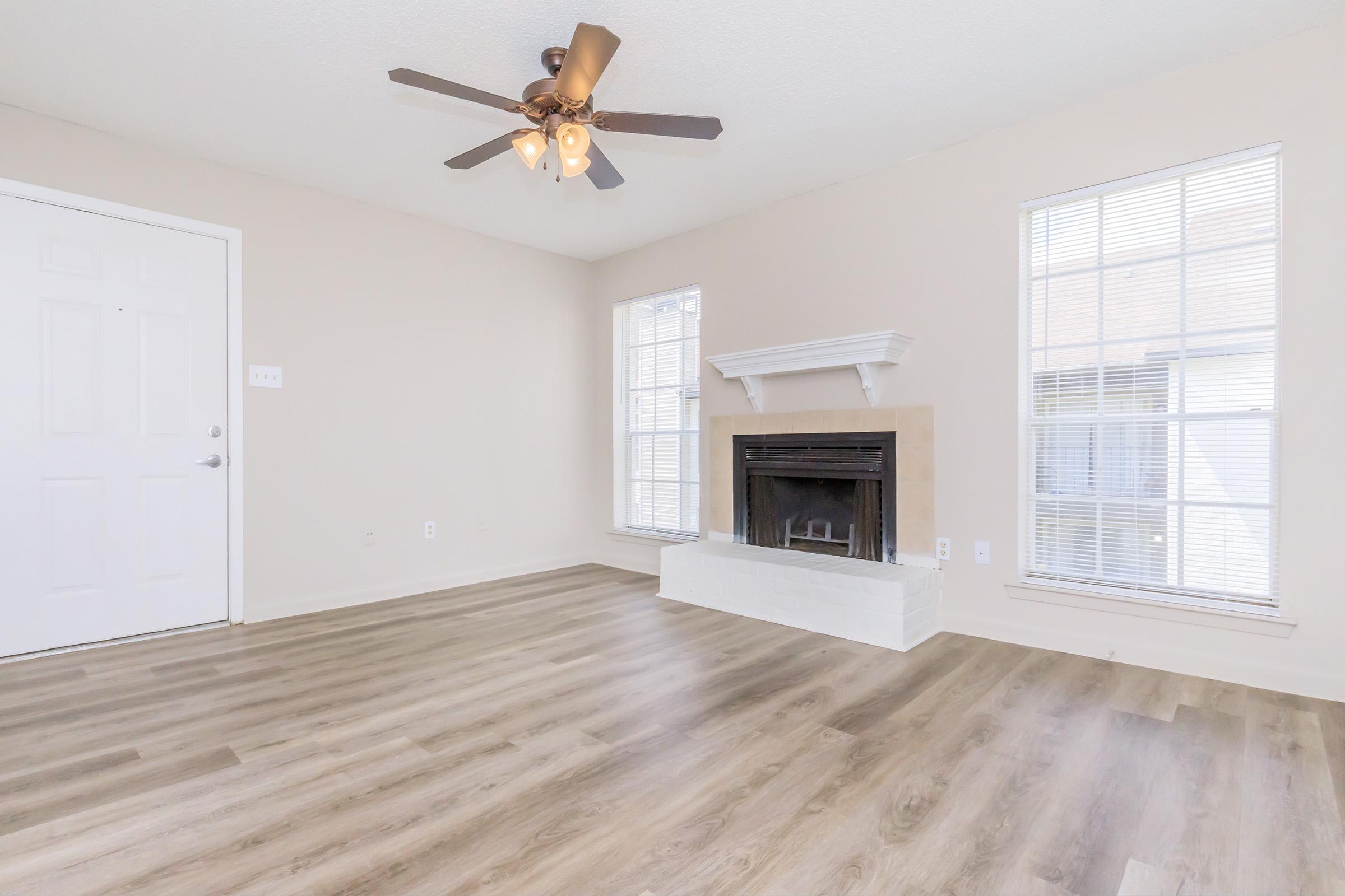 A spacious, well-lit living room featuring a ceiling fan, large windows with blinds, and a white fireplace with a beige surrounding. The floor is covered in light-colored wood laminate, and the walls are painted a soft neutral color, creating a welcoming atmosphere.