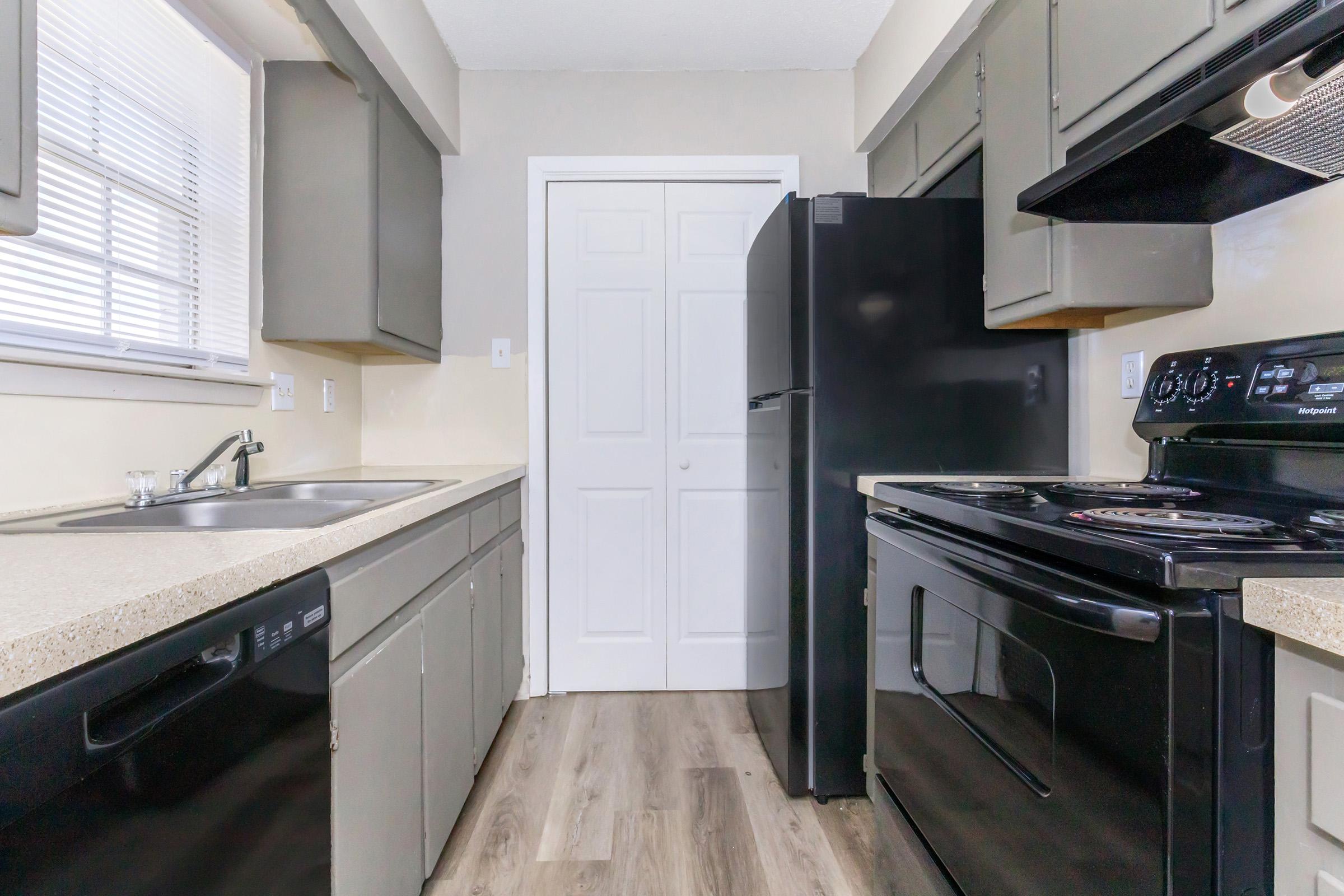 Modern kitchen featuring gray cabinets, a black refrigerator, and a black oven. There is a sink nearby, and the floors are covered in wood-like laminate. Natural light filters in through a window, complementing the neutral wall color. A closed white door is visible at the end of the kitchen area.