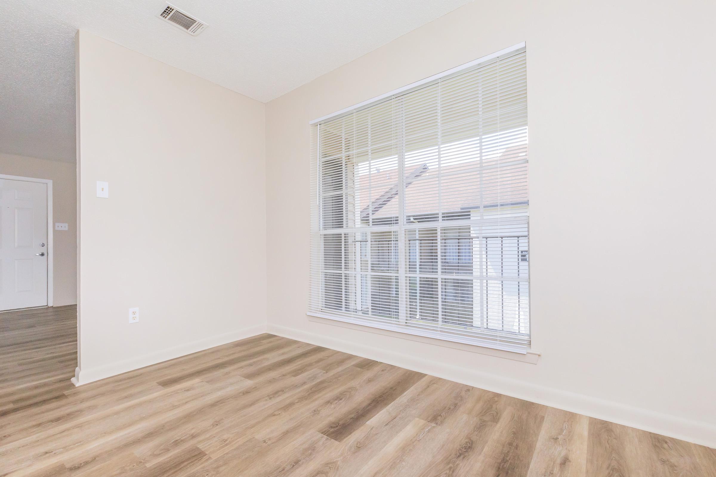 Bright, empty room featuring a large window with blinds, light-colored walls, and laminate flooring. A doorway is visible on the left side, indicating access to another room. The overall atmosphere is spacious and welcoming, ready for furnishing.