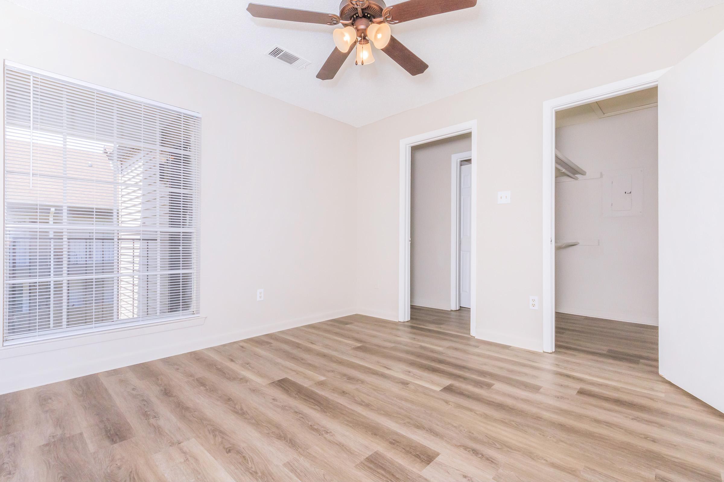 Bright and airy room featuring light-colored walls and wooden flooring. A ceiling fan hangs from the ceiling, and there is a large window with blinds allowing natural light. Two doorways lead to adjacent spaces, one of which appears to be a closet. The overall ambiance is clean and modern.