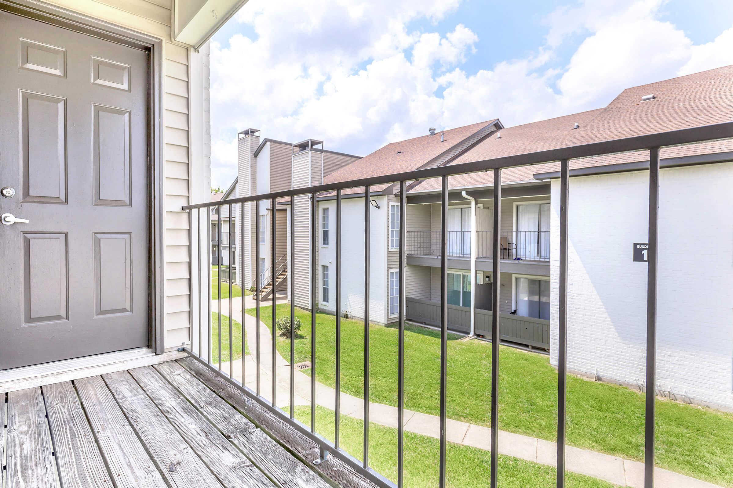 View from a balcony overlooking a landscaped courtyard of an apartment complex. The image features a wooden deck, a metal railing, and multiple buildings with balconies. The sky is partly cloudy, creating a bright atmosphere.