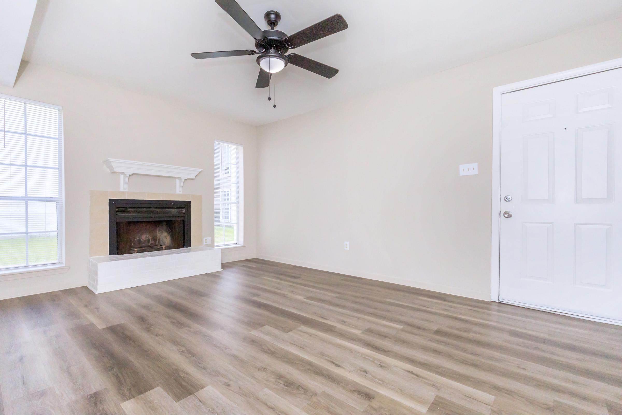 Bright, empty living room featuring light-colored walls and a modern ceiling fan. A white fireplace is centered on the back wall, while large windows on either side allow natural light to enter. The flooring is a warm, wood-like laminate, and a white door leads outside.