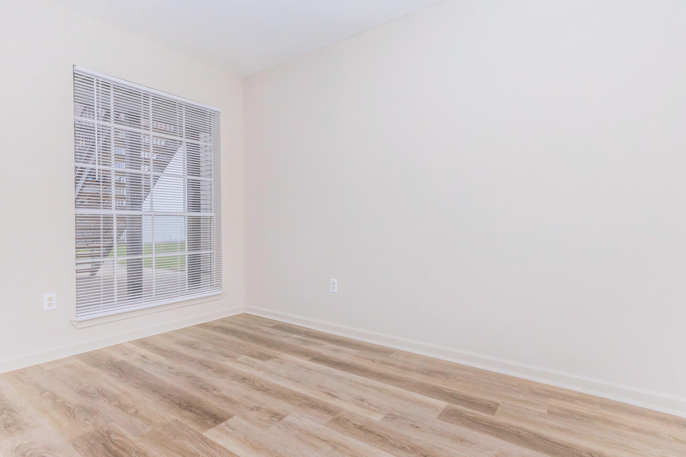 A spacious, empty room with light-colored walls and laminate flooring. A large window with white blinds allows natural light to enter, creating a bright and airy atmosphere. The room is unfurnished, providing a blank canvas for any decor or furniture arrangement.