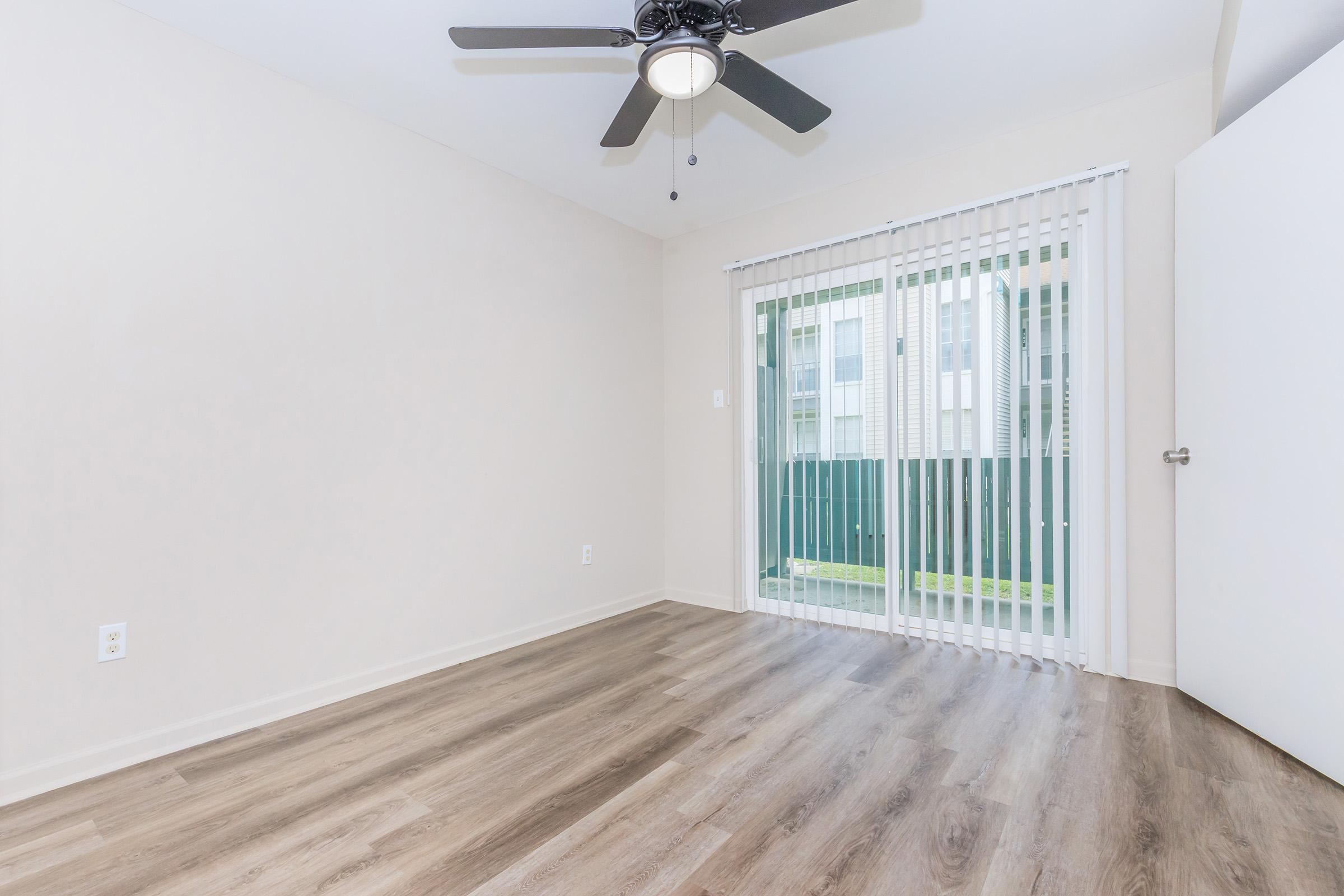An empty room with light-colored walls and wooden flooring. A ceiling fan is visible, and there is a sliding glass door with vertical blinds leading to an outdoor area. The room appears well-lit and spacious, suitable for various uses.