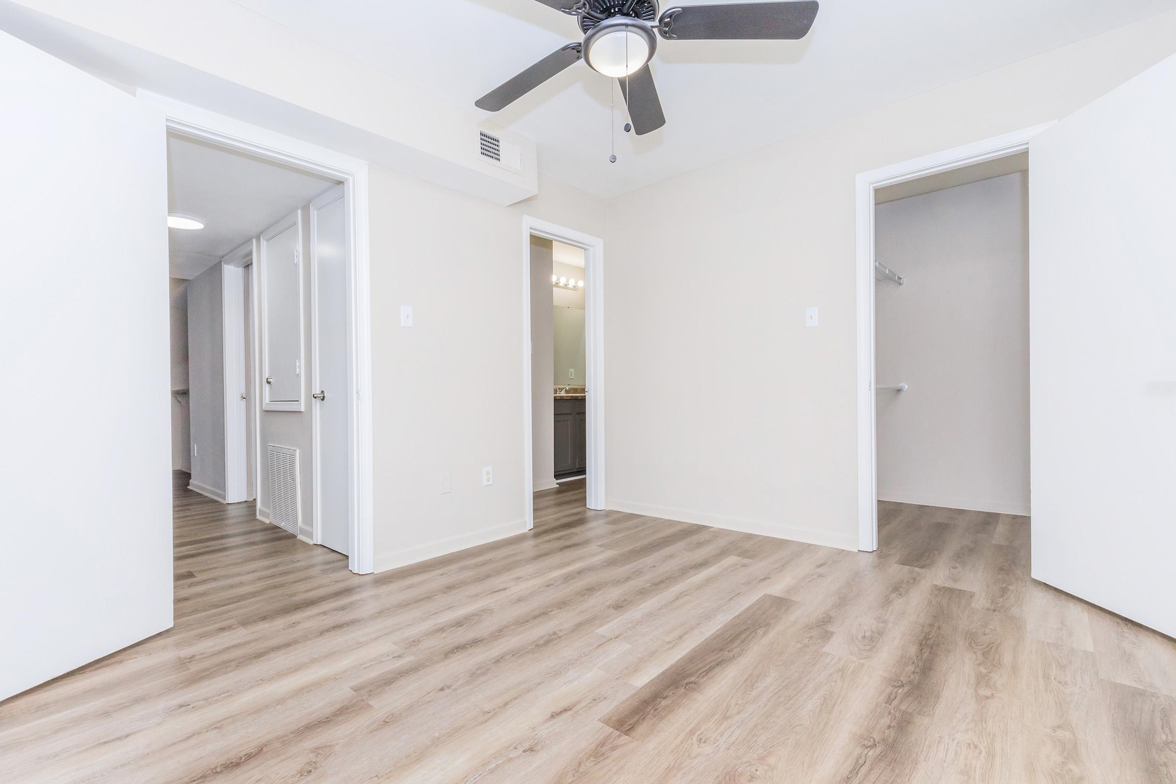 A well-lit interior space featuring light wood flooring, a ceiling fan, and open white doors leading to adjacent rooms. The walls are painted in a neutral color, creating a spacious and inviting atmosphere. The hallway is visible in the background, adding to the room's accessibility.