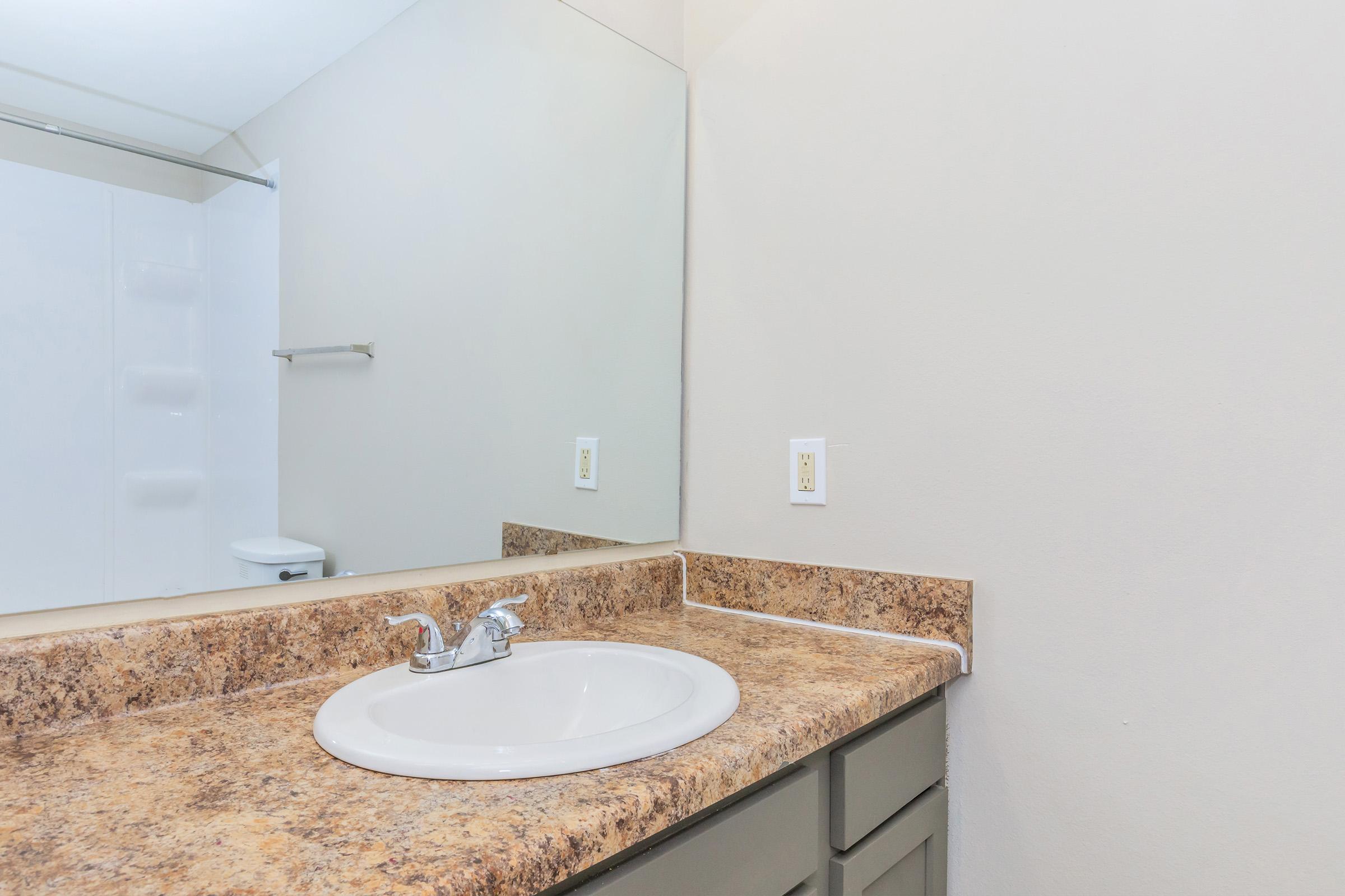 A clean and simple bathroom countertop with a sink, faucet, and a mirror above. The wall is light-colored, and there is a section of a shower area visible in the background. A toilet is also partially visible. The countertop features a speckled brown surface and the cabinetry is a muted green color.