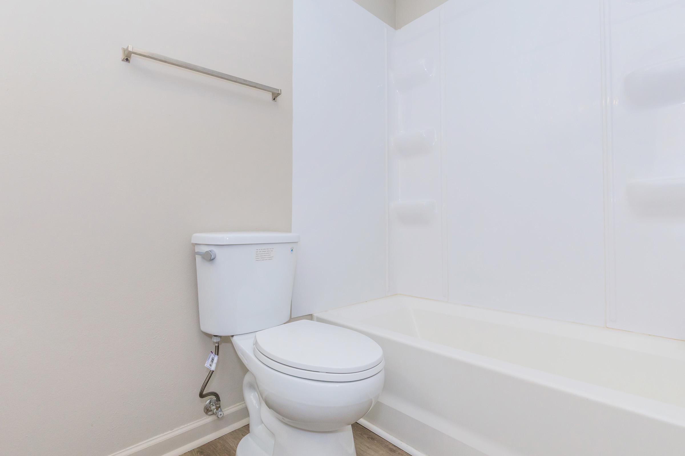 A clean, modern bathroom featuring a white toilet and a bathtub with a shower wall. The walls are a light color, and there is a towel bar mounted on the wall. The overall design is minimalist and well-maintained.
