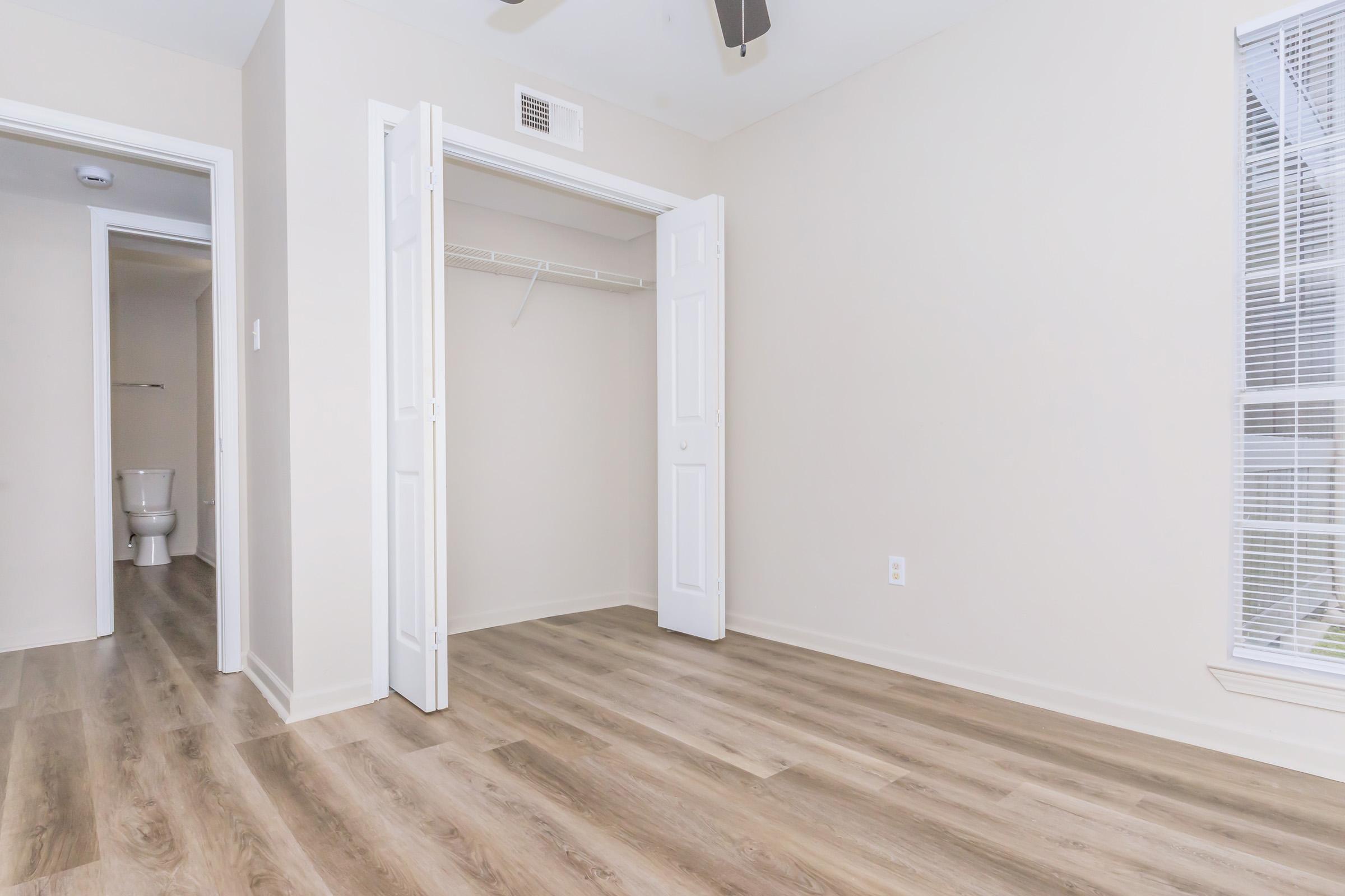 Empty room with light-colored walls and a wooden floor. The room features an open closet with white doors on one side, and a doorway leading to a bathroom visible in the background. Natural light comes in through a window, adding brightness to the space.