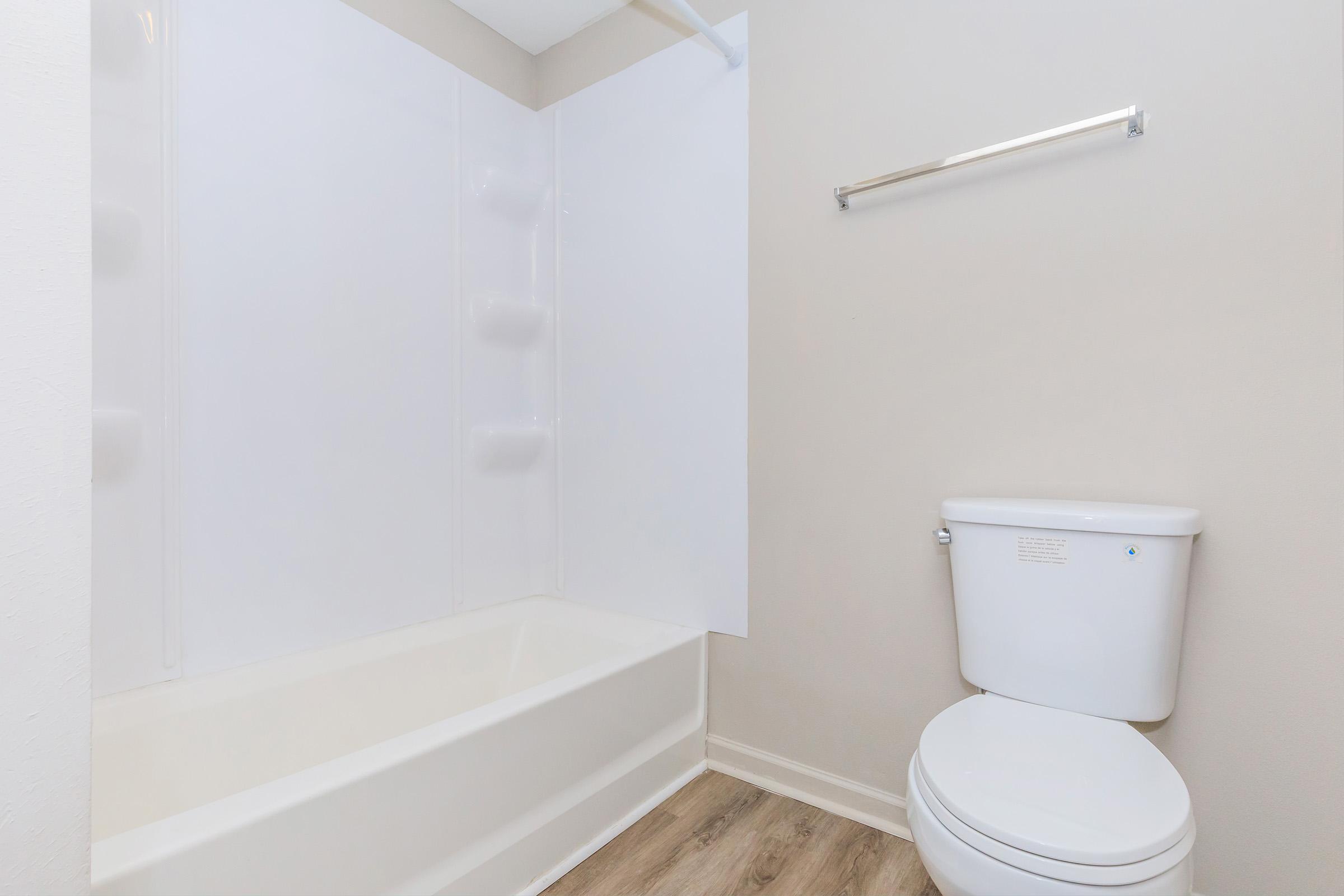 A clean, modern bathroom featuring a white bathtub, a wall-mounted towel rack, and a white toilet. The walls are painted in a light color, and the flooring is a wood-like laminate, creating a bright and minimalistic aesthetic.