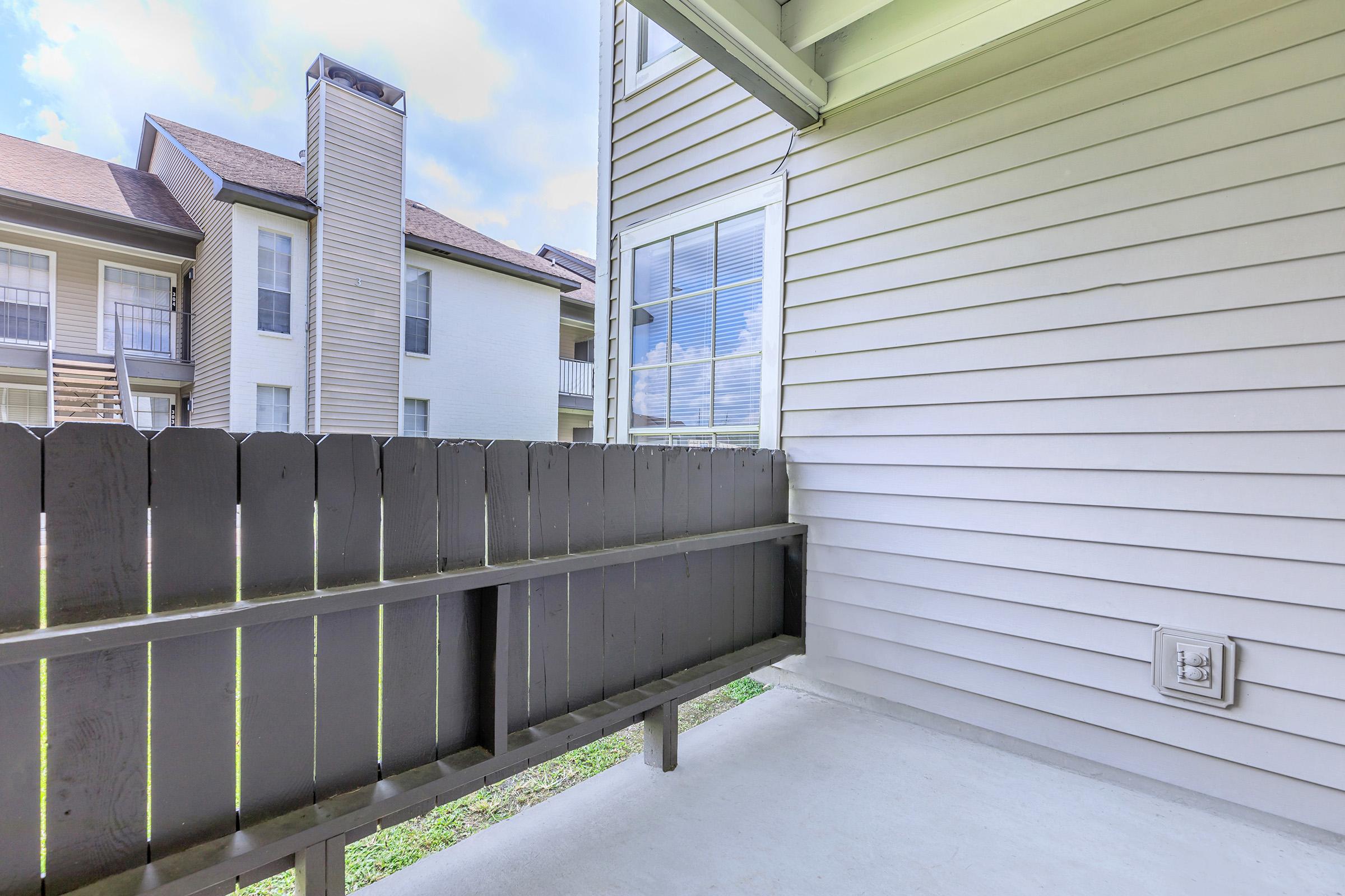 A view of a balcony or patio area featuring a wooden fence, with neighboring apartment buildings visible in the background. The walls are light-colored, and the sky is partly cloudy. The space is clean and empty, suggesting it is ready for outdoor furniture or personal use.