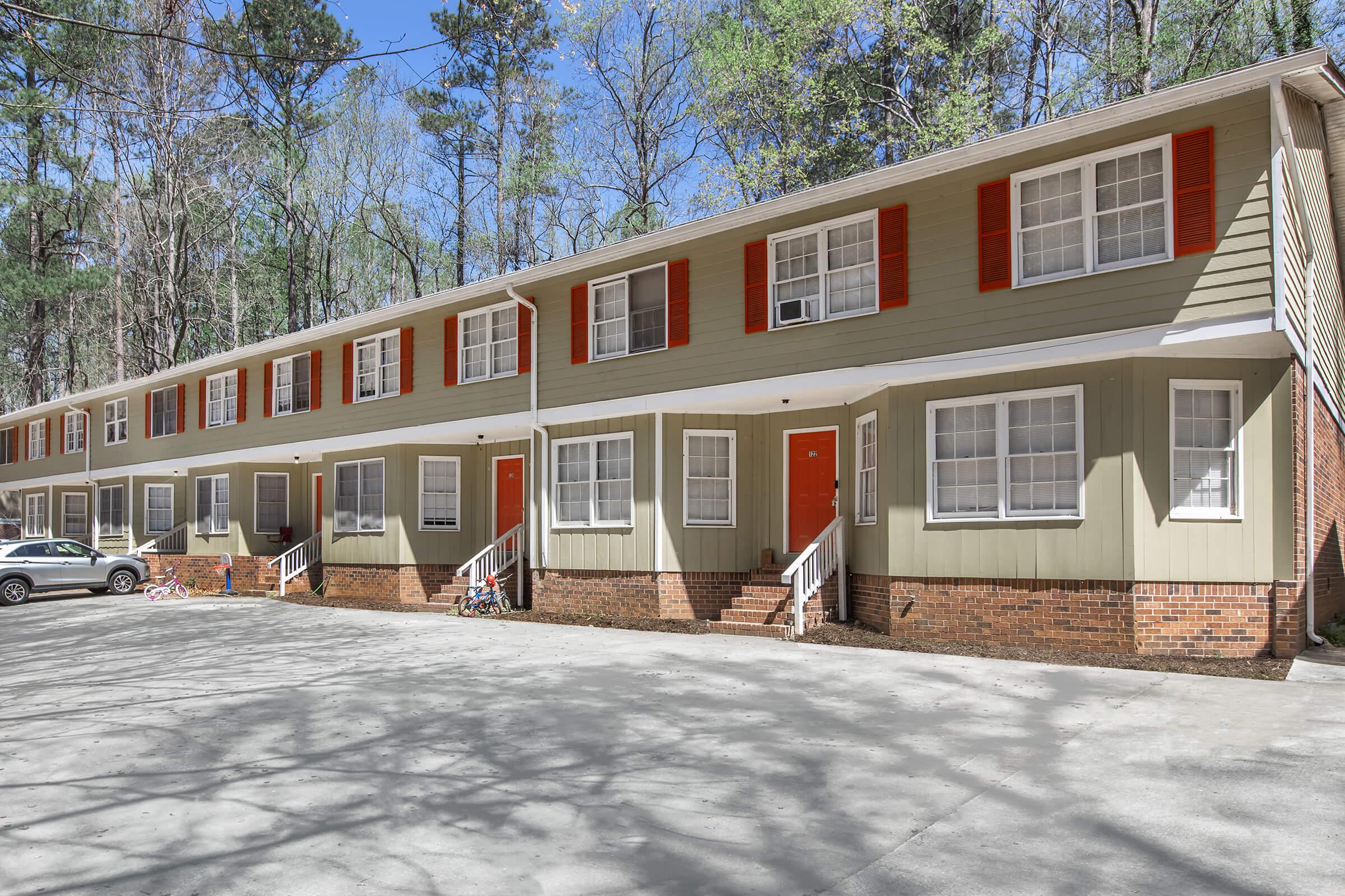 A row of residential buildings with two stories, featuring light green siding and red shutters. The front doors are orange, and there are small staircases leading to each entrance. Surrounding trees indicate a wooded area, and a parking space is visible in front of the buildings.