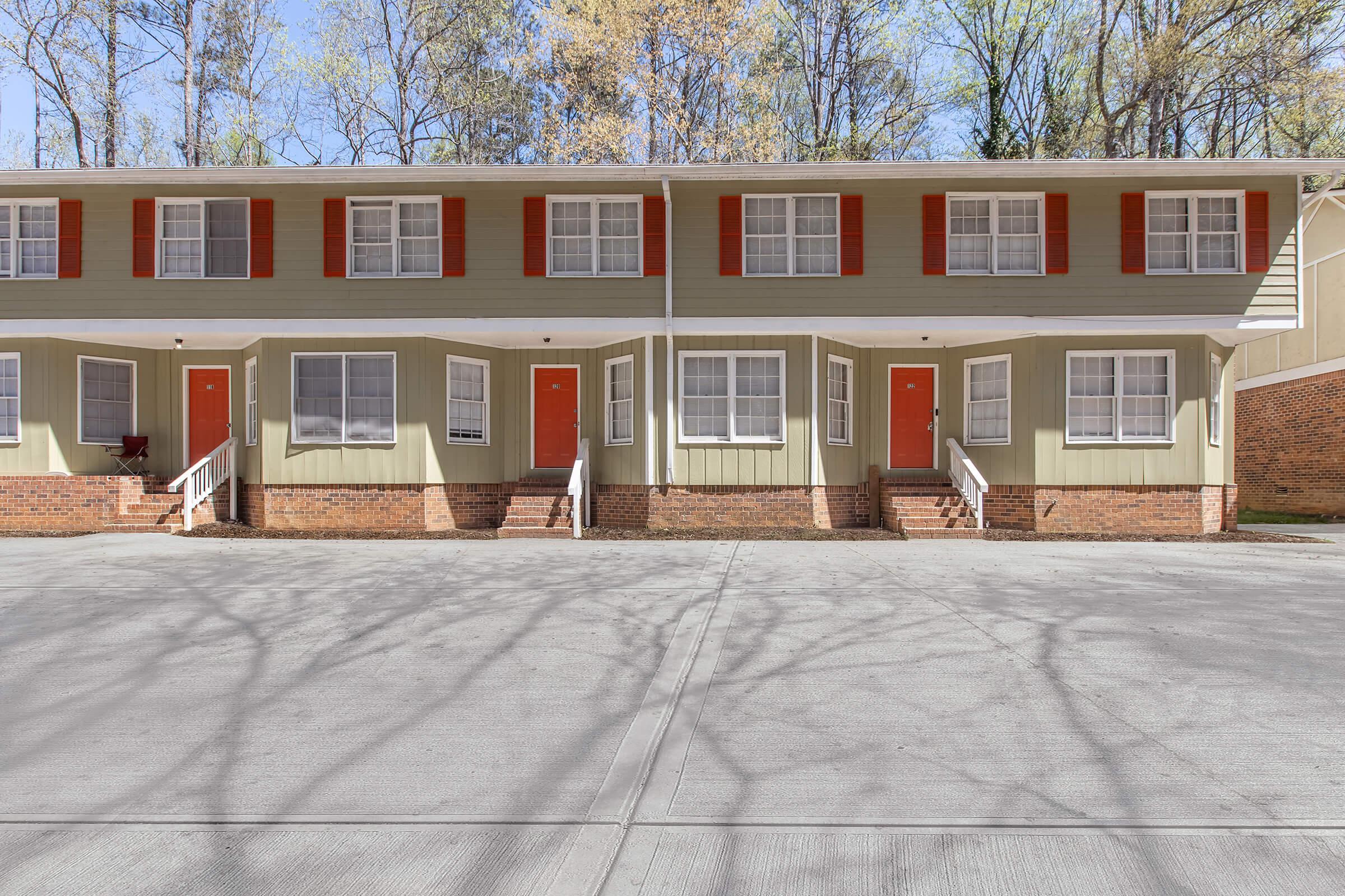 A row of two-story townhouses with light green exteriors and red shutters. Each unit features a front door painted orange and has windows with white trim. The setting includes a paved area in front and trees in the background, indicating a suburban environment.