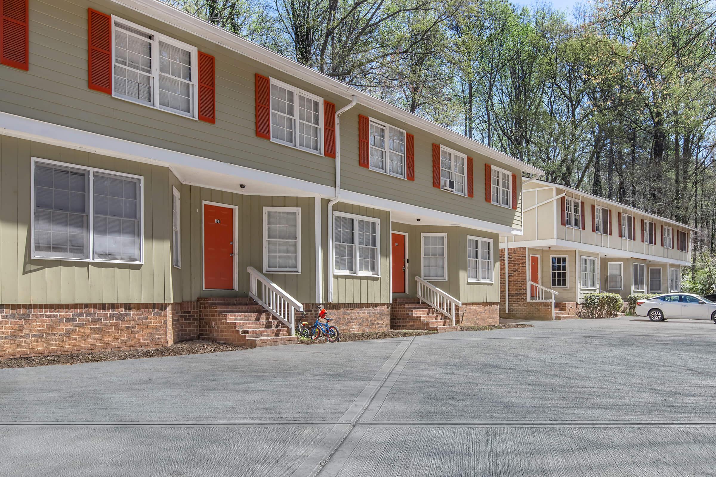 A row of multi-family residential units with green and beige siding, featuring red shutters. Each unit has a front porch with steps leading to the entrance. The area is surrounded by trees, and a small child is playing with a bicycle in front of one of the units. The driveway is paved with a few parked cars.