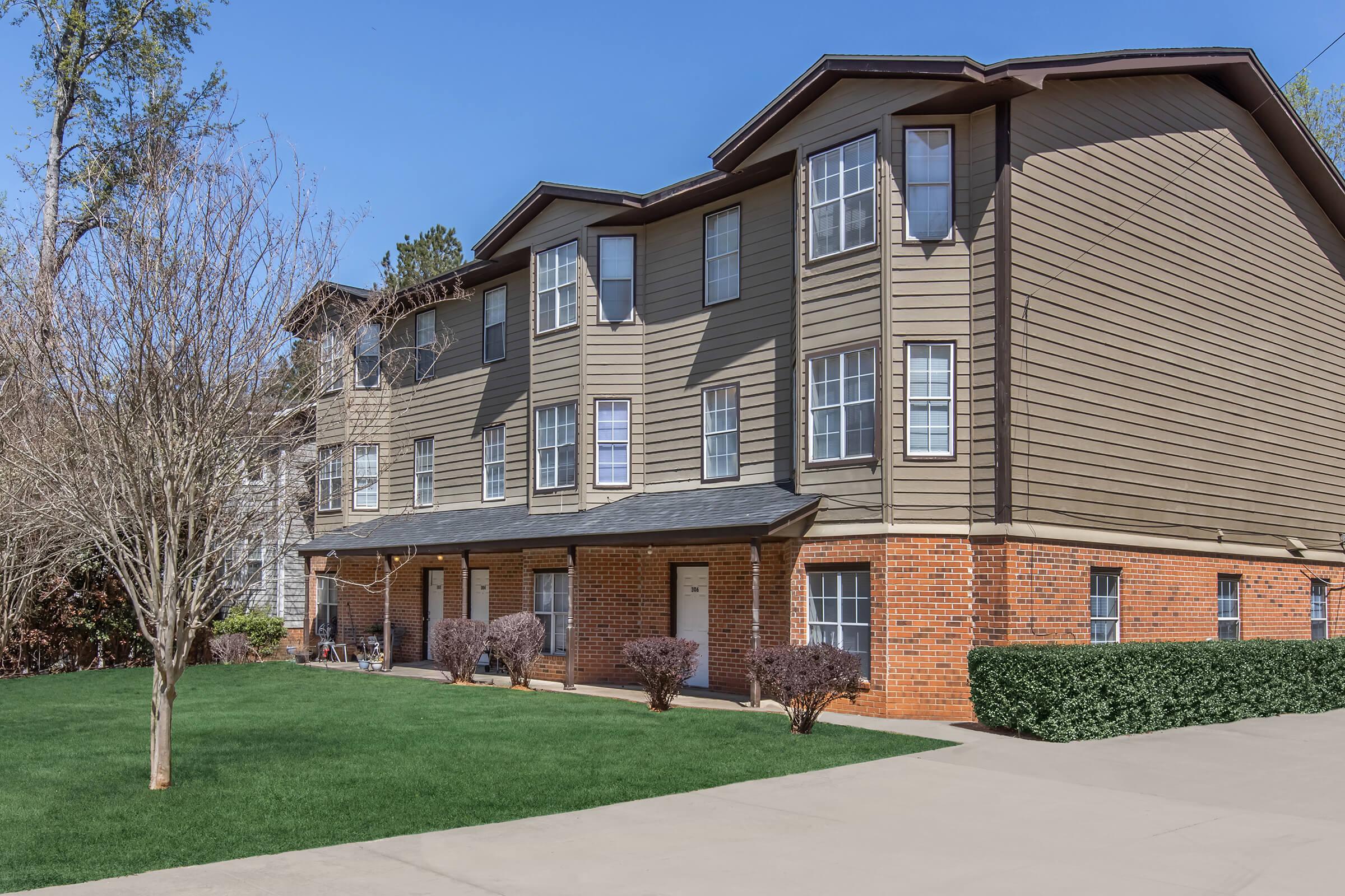 A three-story apartment building with a combination of brown siding and brick accents. The facade features several windows and front doors on the lower level. In front, there's a well-maintained green lawn, with small shrubs along the building's base, set against a clear blue sky.