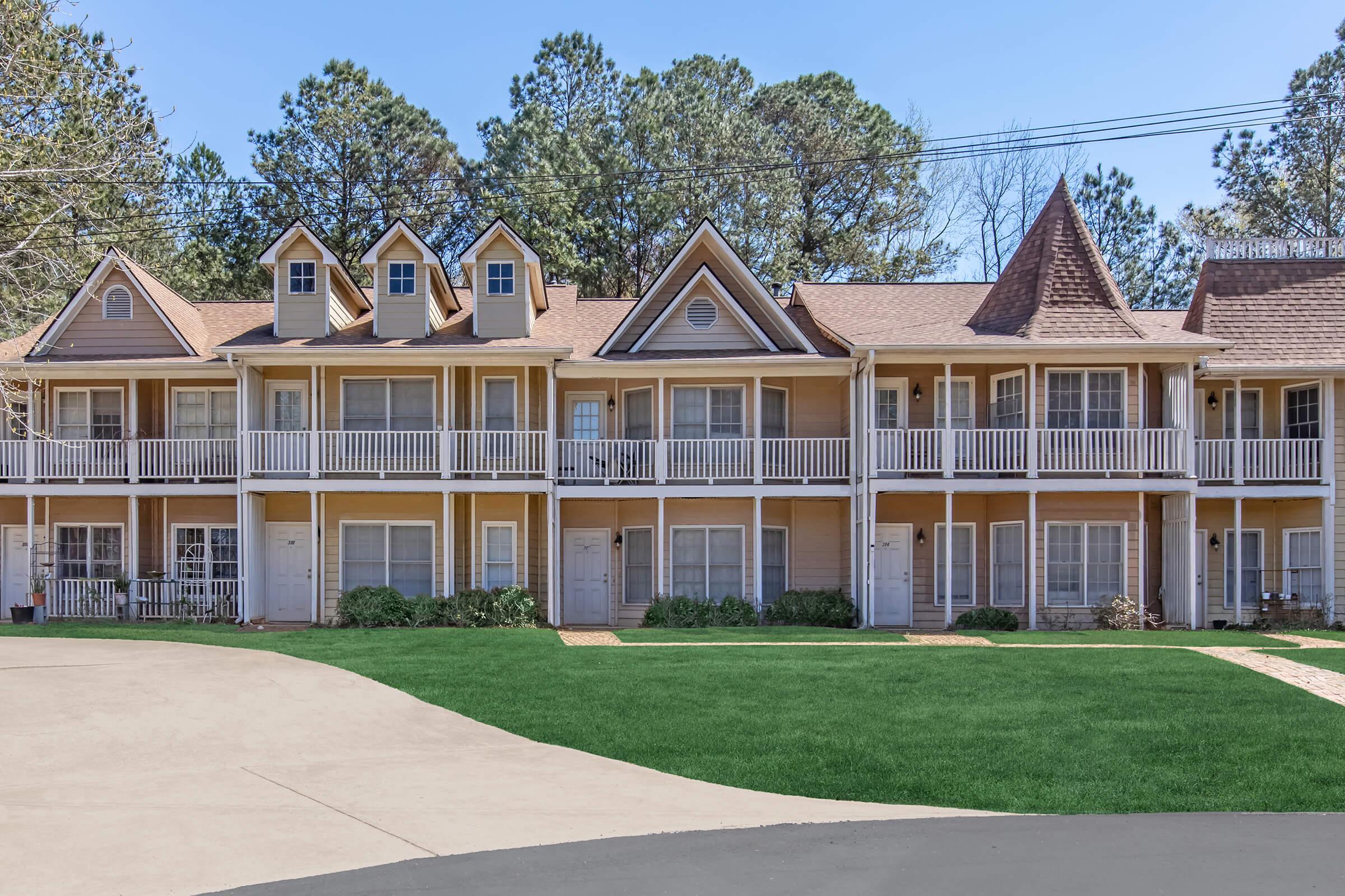 A row of two-story, light brown townhouses with balconies, set against a backdrop of tall trees and blue skies. The grassy area in front is well-maintained, and the driveways are paved, contributing to a clean, inviting appearance.