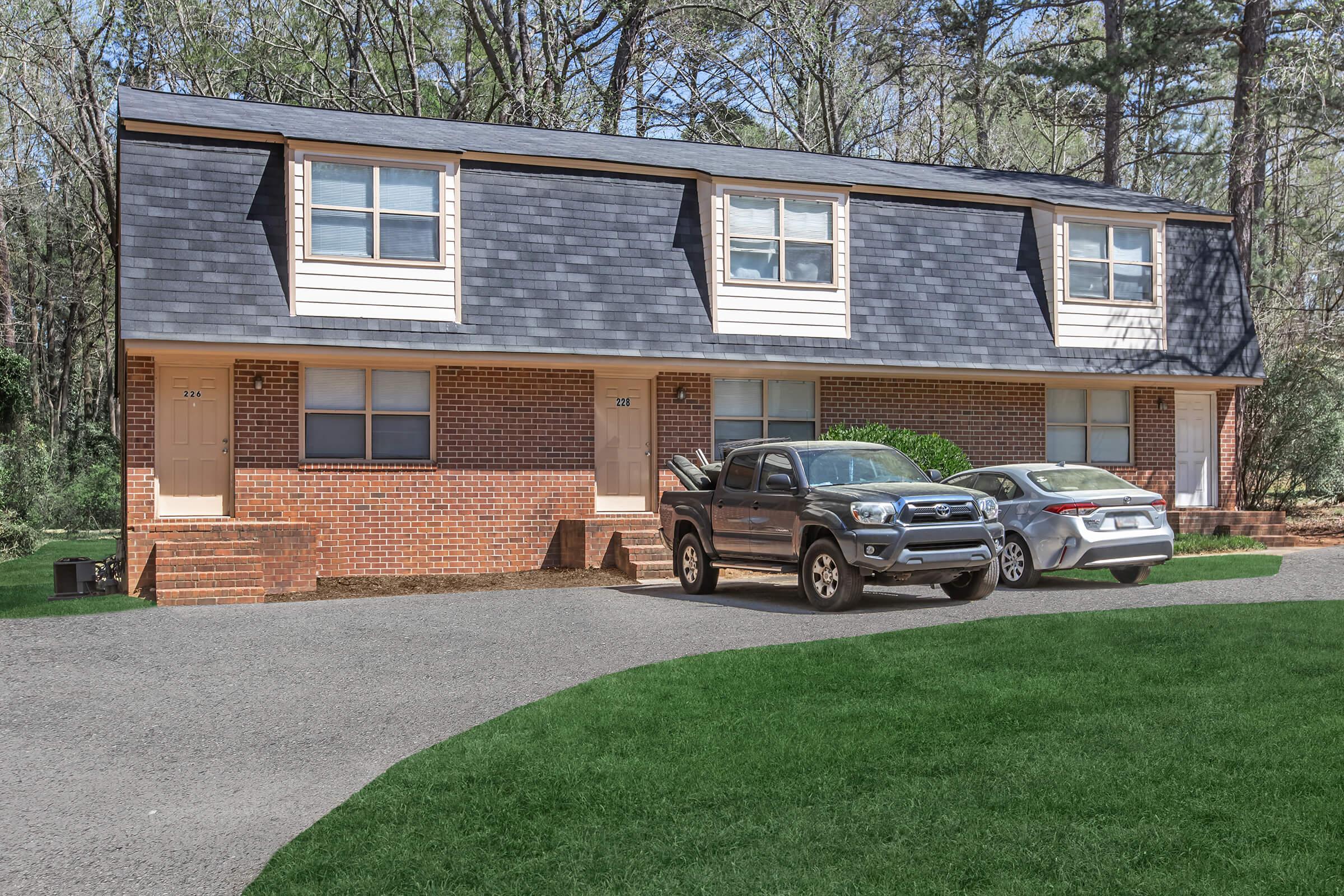 A two-story brick duplex surrounded by trees, with two parked cars in front. The building features two entrances on each side, with beige siding on the upper level and dark shingles on the roof. The driveway is paved, and the lawn is well-maintained with green grass.