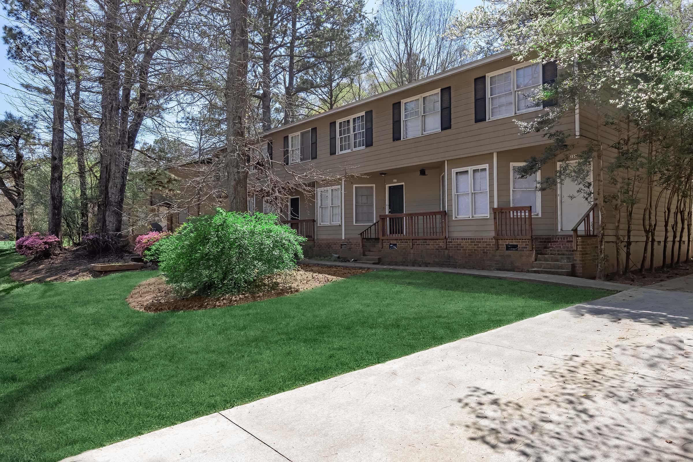 A two-story beige house with dark shutters, surrounded by tall trees and blooming shrubs. The front yard features a well-maintained green lawn and a small landscaped area. The driveway leads up to the house, providing easy access. Natural light illuminates the exterior, creating a welcoming atmosphere.