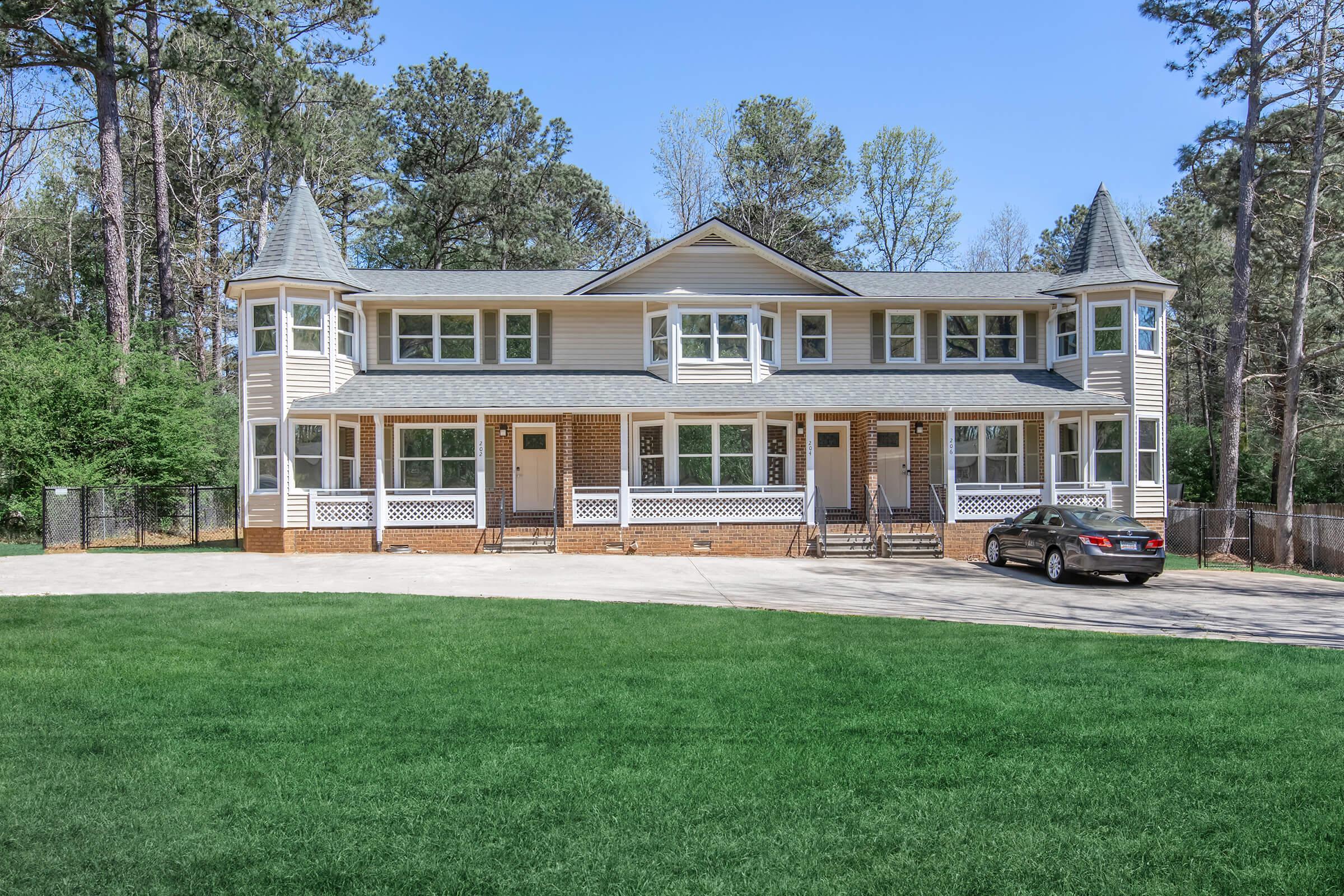 A two-story residential building featuring multiple entrances and a spacious yard. The exterior is predominantly beige with dark gray roofing. Green grass surrounds the building, and a car is parked in the driveway. Tall trees can be seen in the background, adding to the property’s serene atmosphere.