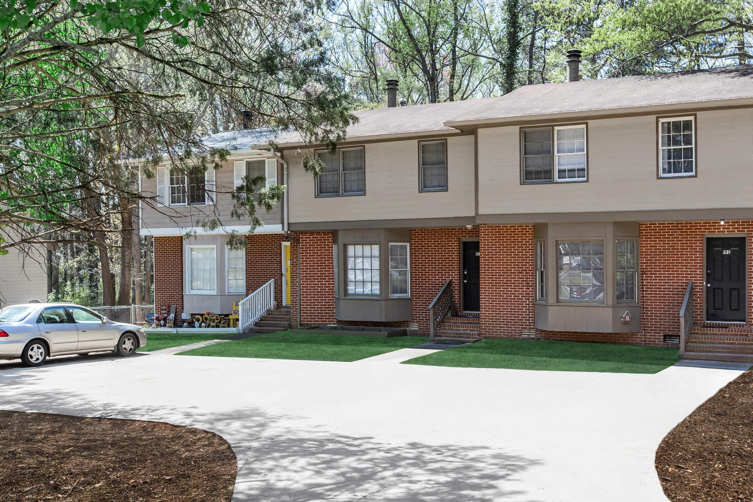 Two adjacent townhouses with brick and siding exteriors in a wooded area. Each unit has a front porch, with one unit having a white staircase. A silver car is parked in the driveway, which is lined with freshly cut grass and surrounded by trees. The scene is bright and welcoming.