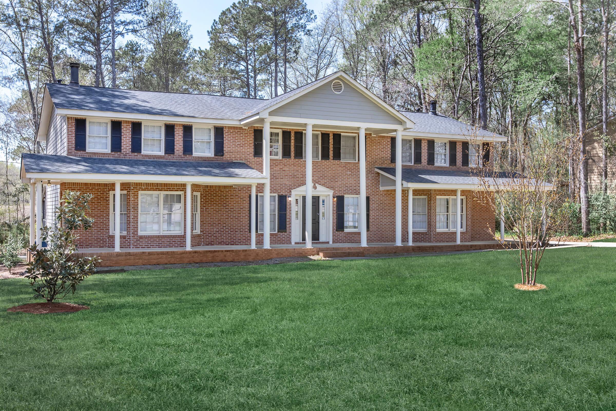 A large, two-story brick house with white trim and multiple windows, situated on a green lawn surrounded by trees. The house features a symmetrical design with a central entrance and a covered porch. Sunlight illuminates the exterior, highlighting the well-maintained landscaping.