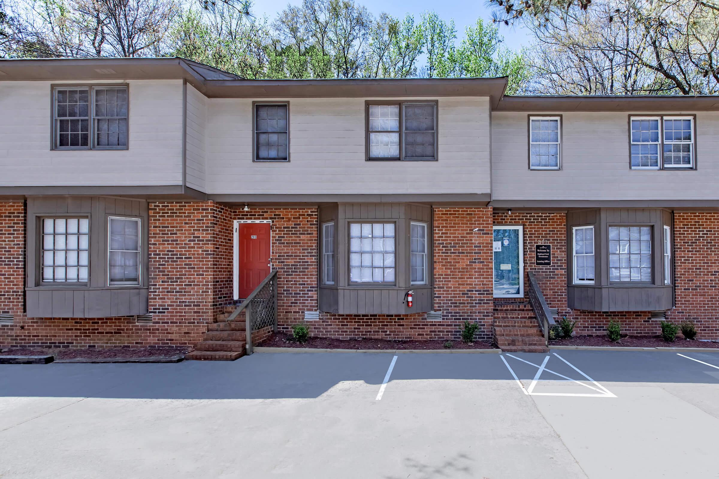 Exterior view of a multi-unit townhouse building. The structure features a beige facade with dark brown accents and a combination of windows. A single red door stands out at the center, and there are two parking spaces visible in front. Lush trees provide greenery in the background.