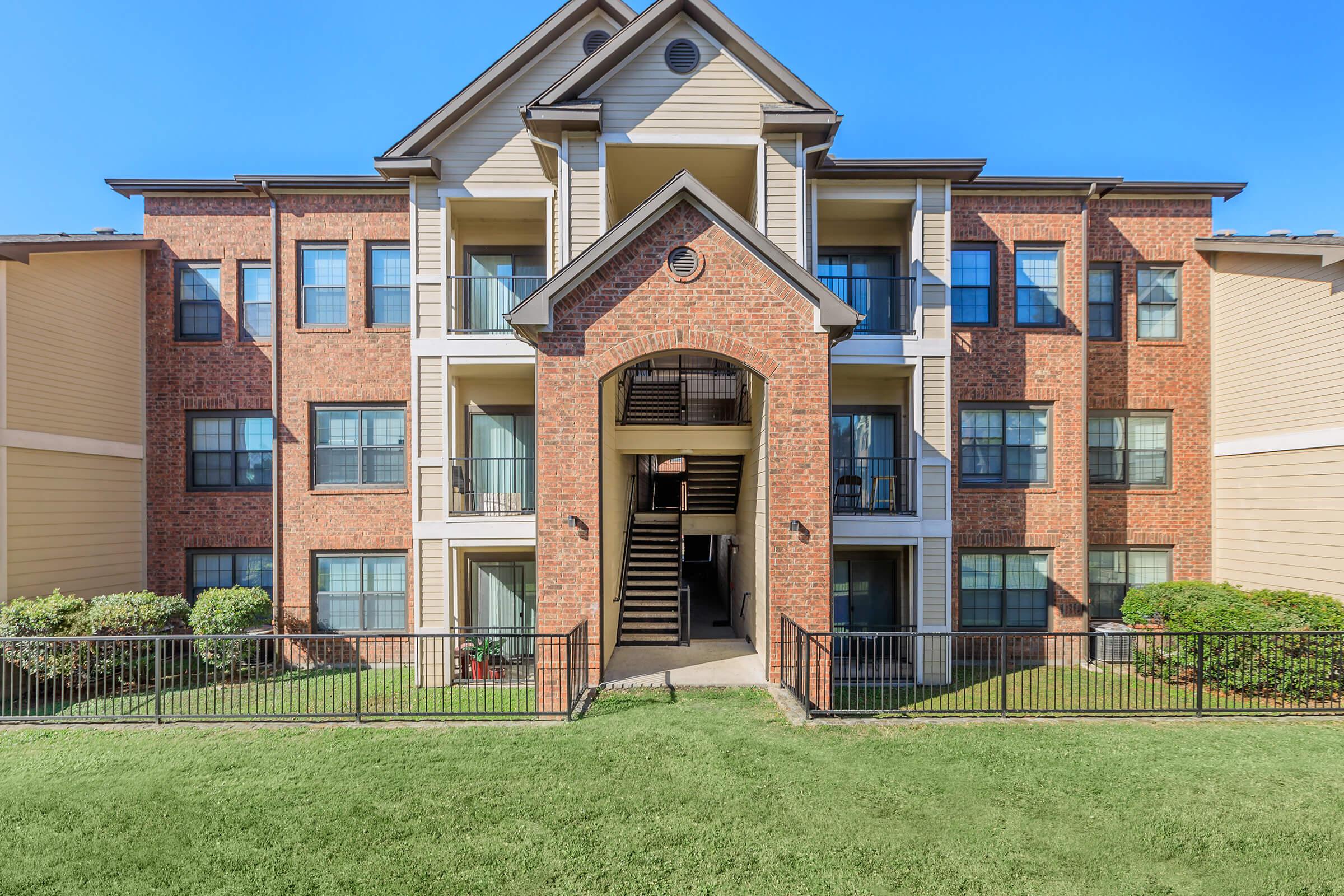 A three-story apartment building with a brick exterior and white accents. The entrance features a covered porch and stairs leading to the upper floors. Surrounding the building are well-maintained lawns and bushes, with clear blue skies above.