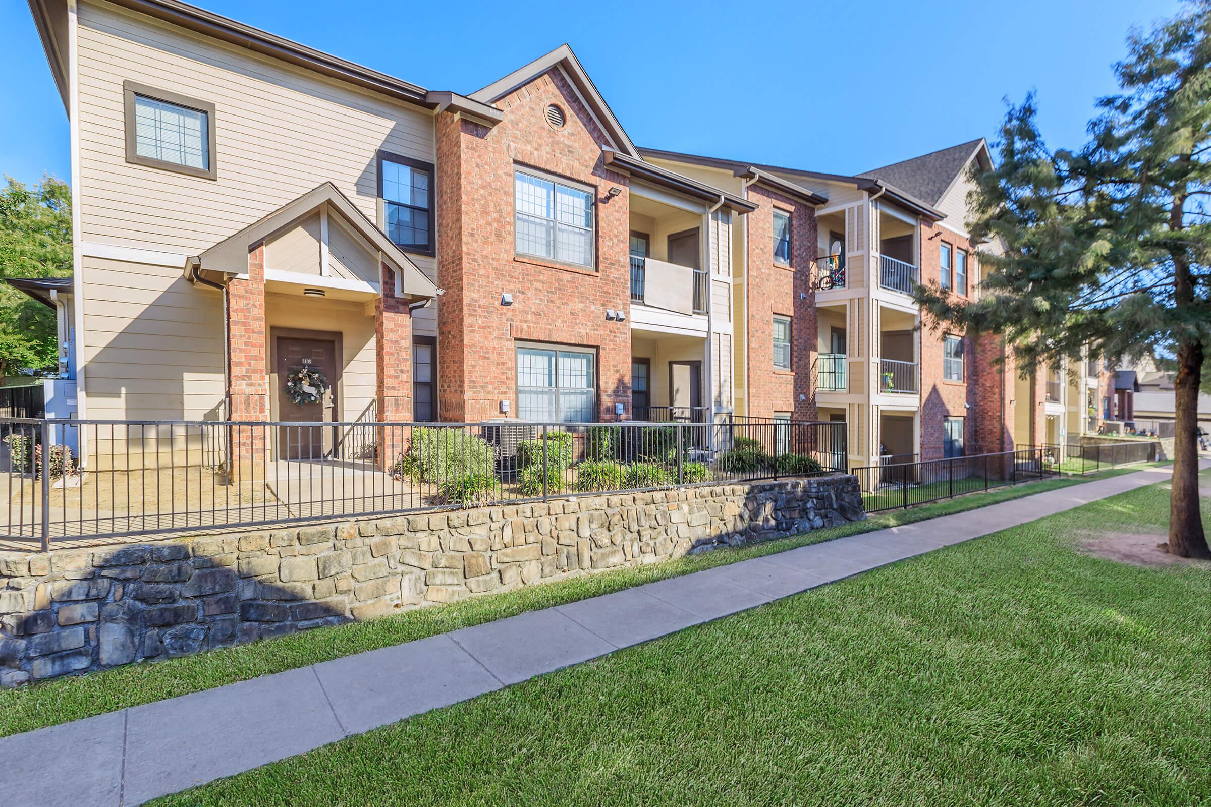 A multi-story apartment building featuring a combination of brick and siding exteriors. The front has a small porch with a decorative wreath, surrounded by landscaped greenery. A walkway leads to the entrance, and balconies are visible on the upper floors.