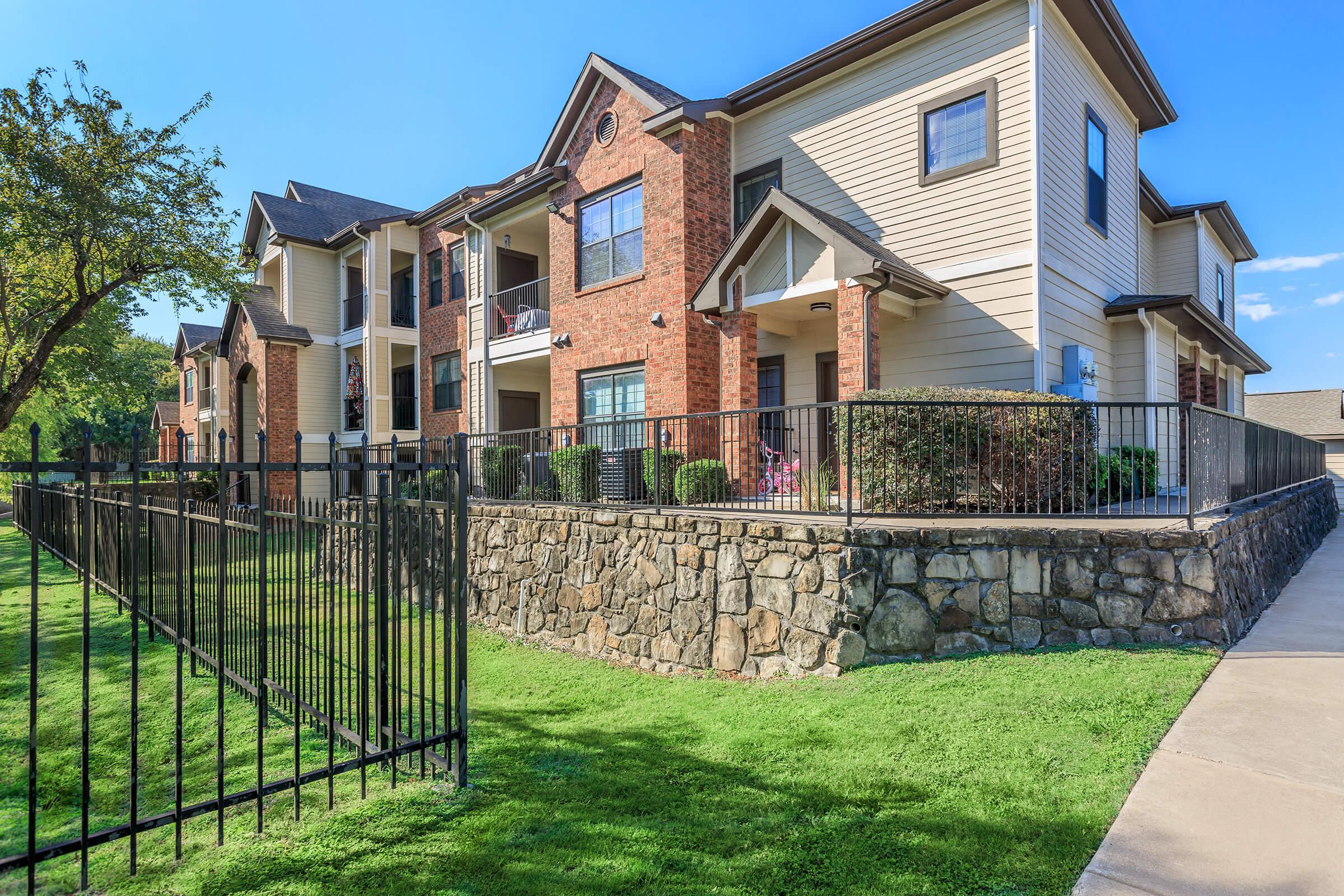 A multi-story apartment building with a mix of brick and siding exteriors. The complex features a stone wall and a well-maintained lawn, surrounded by a black iron fence. A clear blue sky is visible above, and there are balconies with plants on the upper levels.